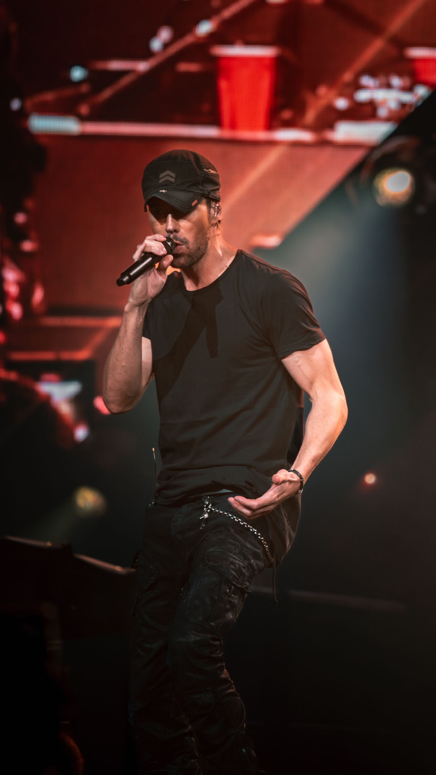 Stage performer in all black outfit singing into microphone with red lighting and cups visible in background.