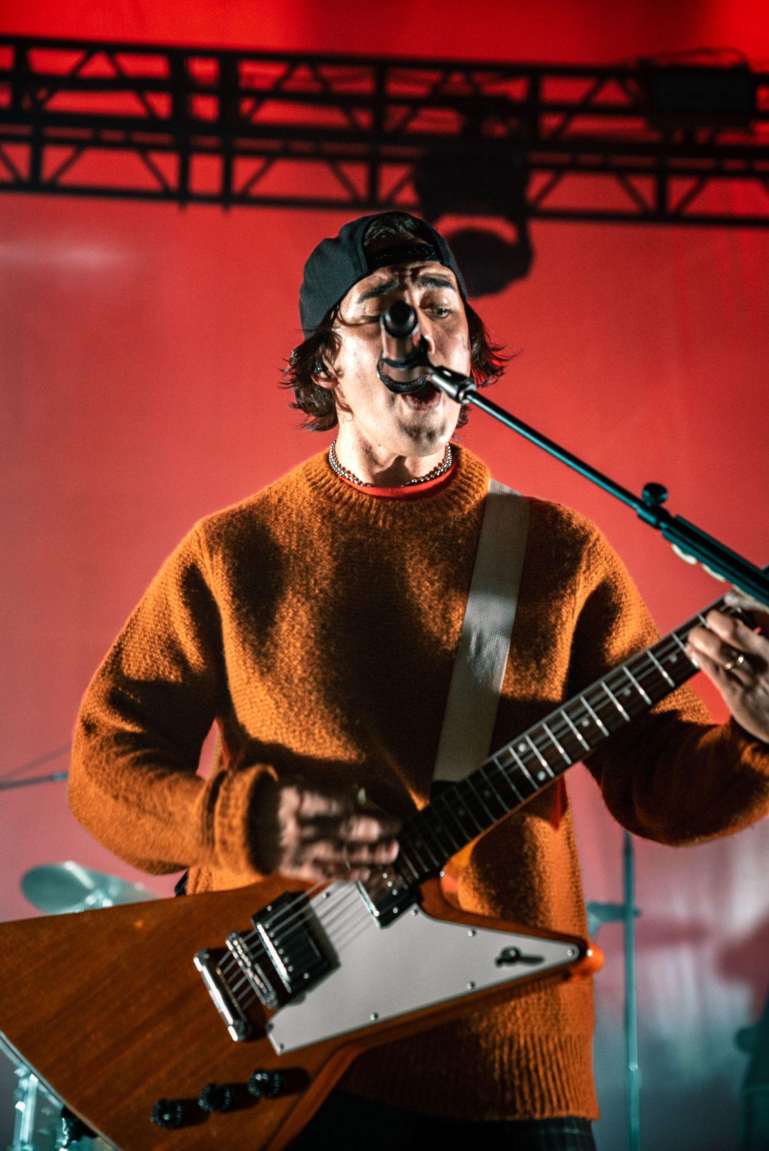 Guitarist wearing orange sweater plays electric guitar under red stage lights.