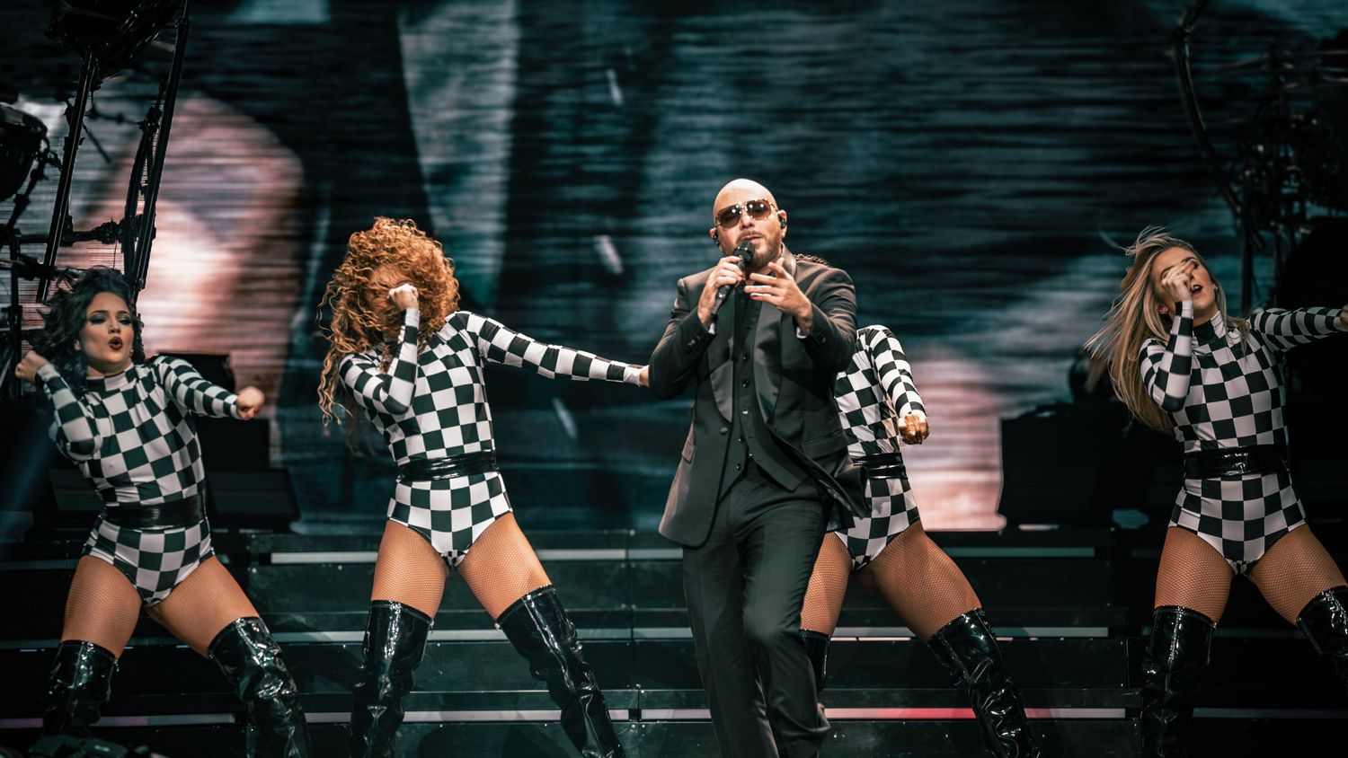 Concert performance featuring dancers in black and white checkered outfits alongside performer in dark suit.