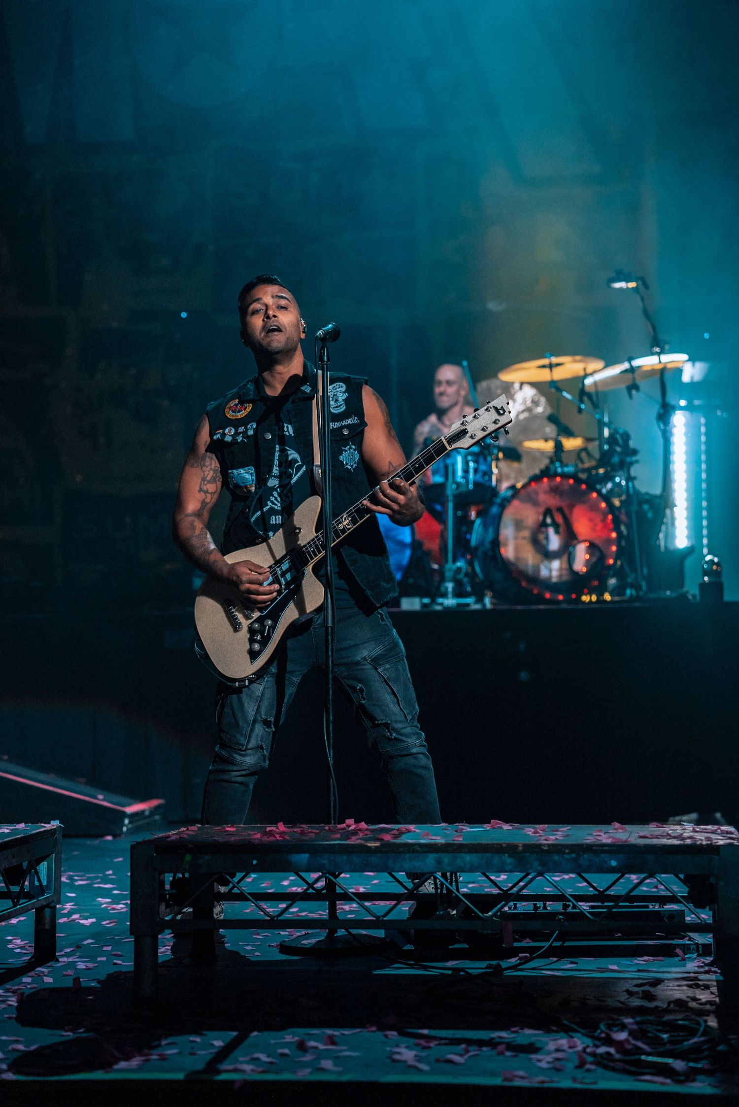 Musician performs on elevated platform with blue stage lighting and drum kit in background.