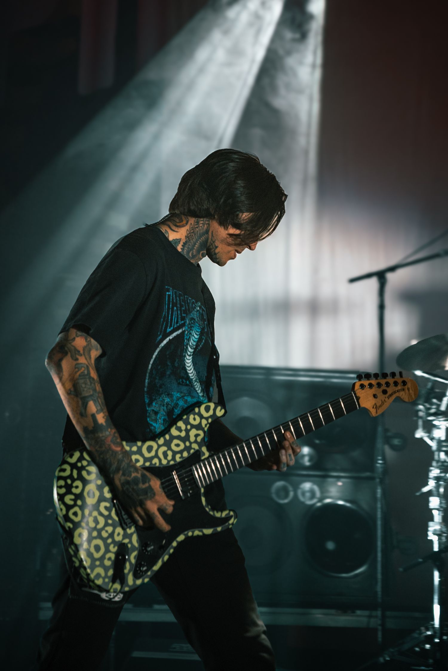 Guitarist with tattoos plays electric guitar under blue stage lighting wearing patterned shirt.