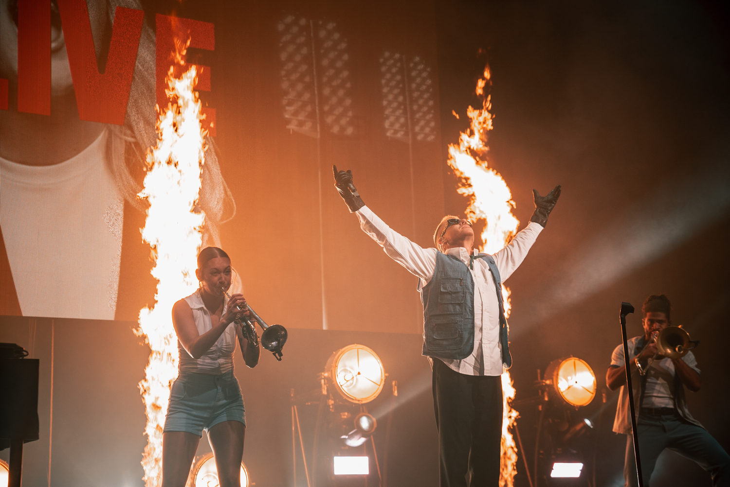 Concert performers on stage with dramatic fire effects and bright spotlights in the background.