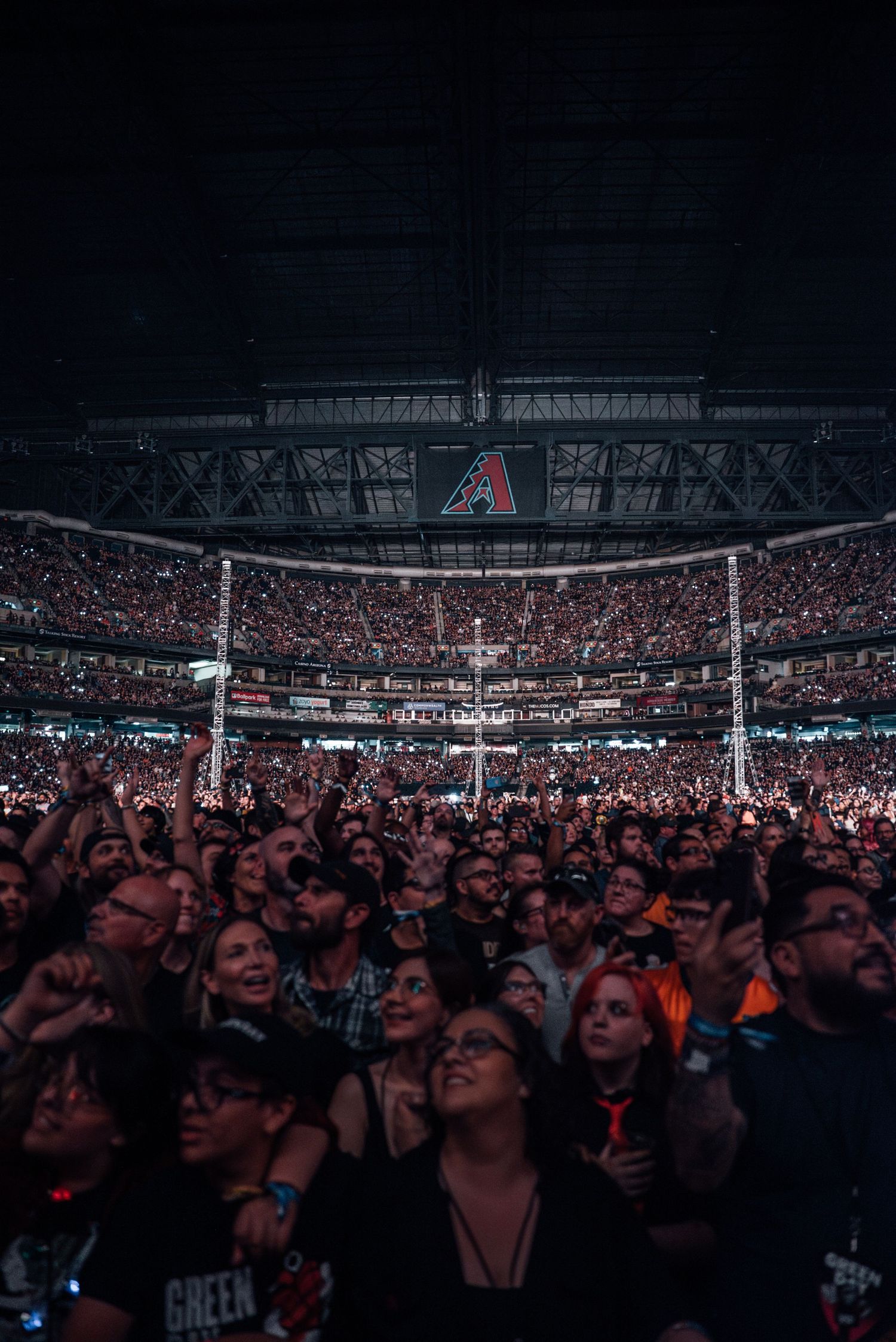A massive crowd fills a stadium arena during a concert with the Arizona Diamondbacks logo visible overhead.