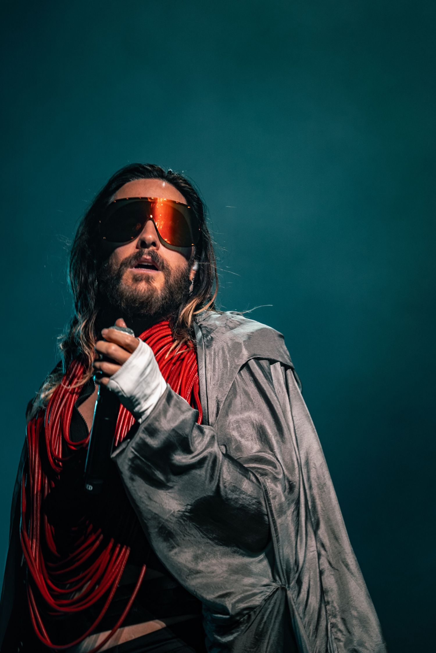 An artist in futuristic sunglasses and grey jacket poses against a teal background.