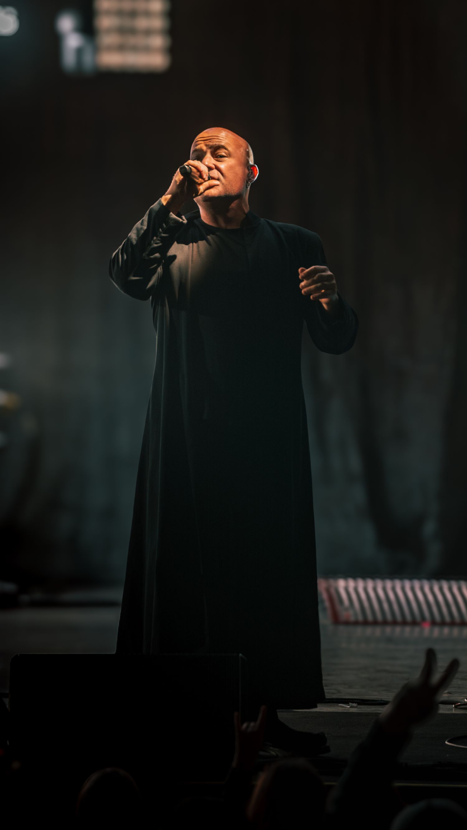 Performer in black attire singing emotively on stage during dramatic concert performance.