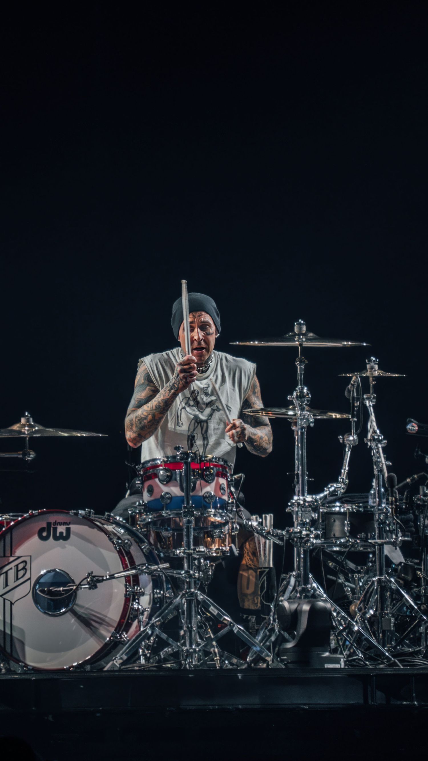 Drummer sits at extensive drum kit setup illuminated against dark stage background.