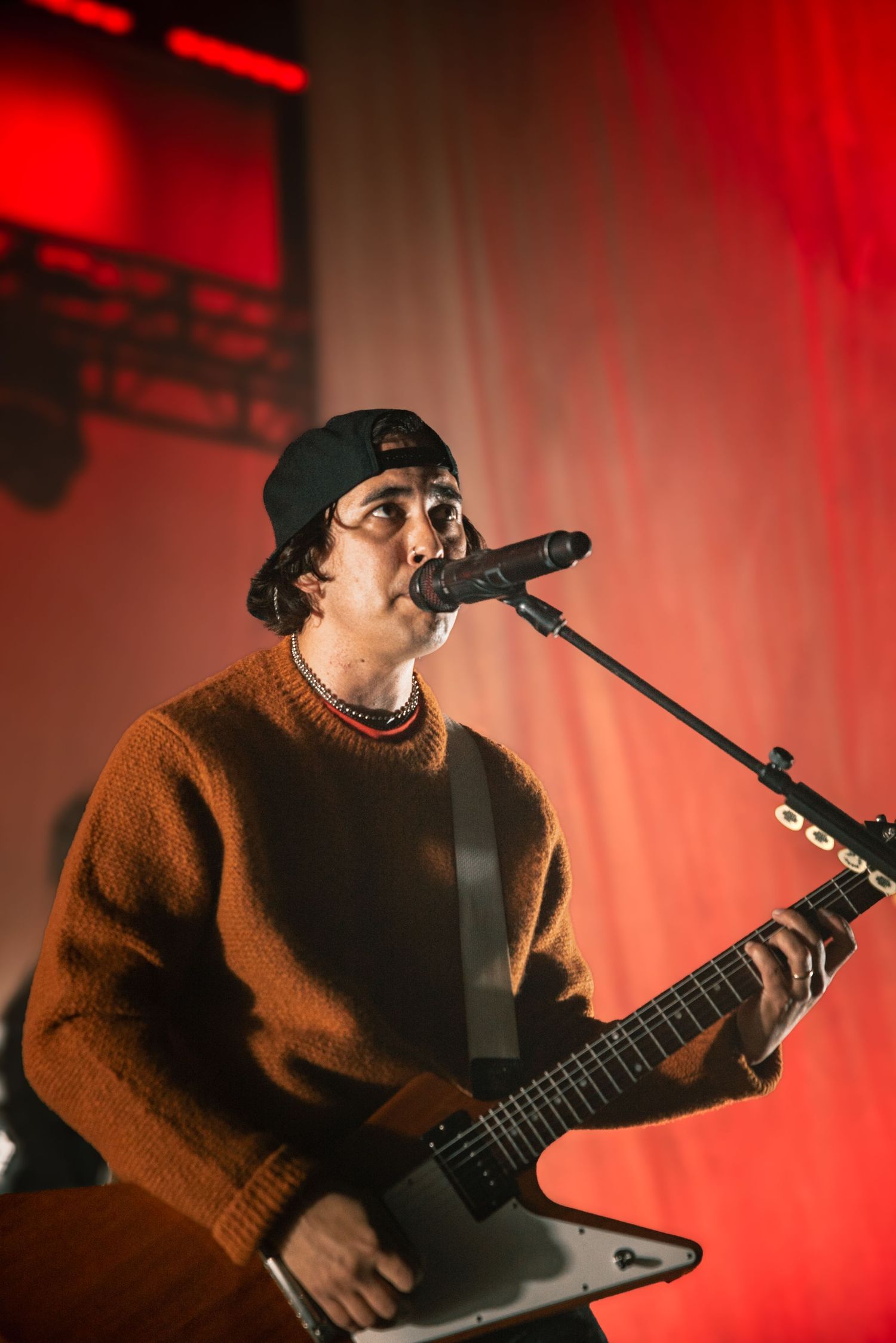 Musician in orange sweater and backwards cap performs with electric guitar against red-lit backdrop.