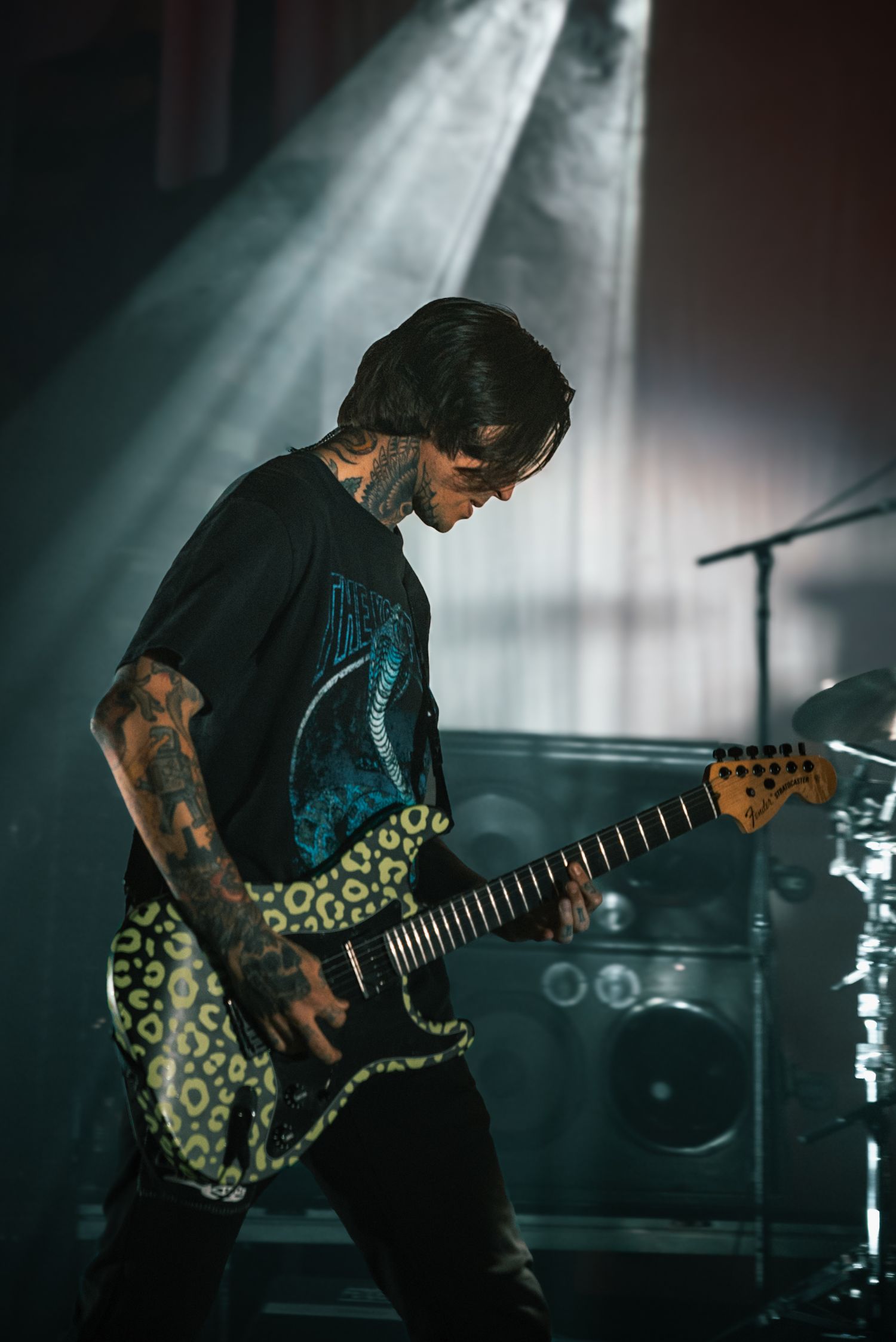 Rock guitarist performs with tattooed arms and patterned shirt under dramatic stage lighting.