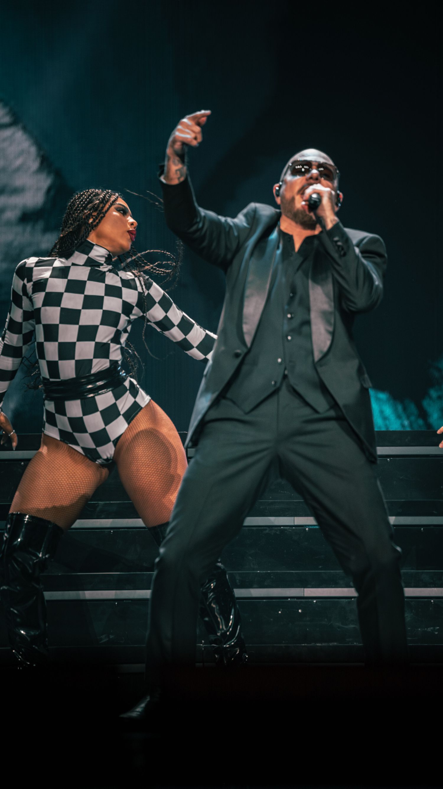 Dynamic stage duet with performer in suit and dancer in checkered bodysuit pointing upward.