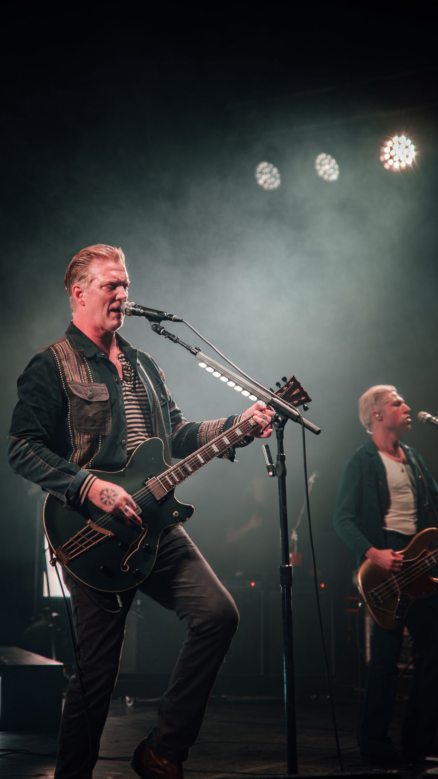Band members perform on smoky stage with dramatic lighting while playing guitars during rock concert.