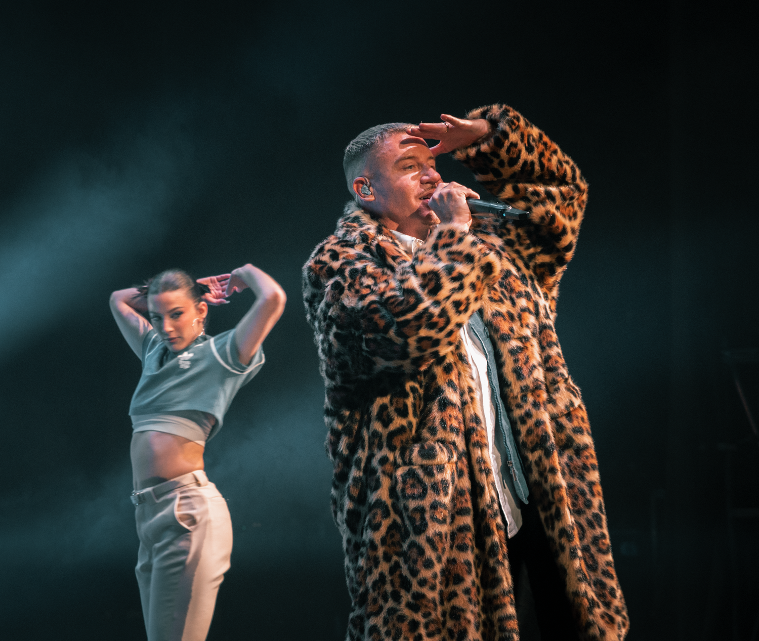 Dancers perform on stage with one wearing a striking leopard print coat under atmospheric lighting.