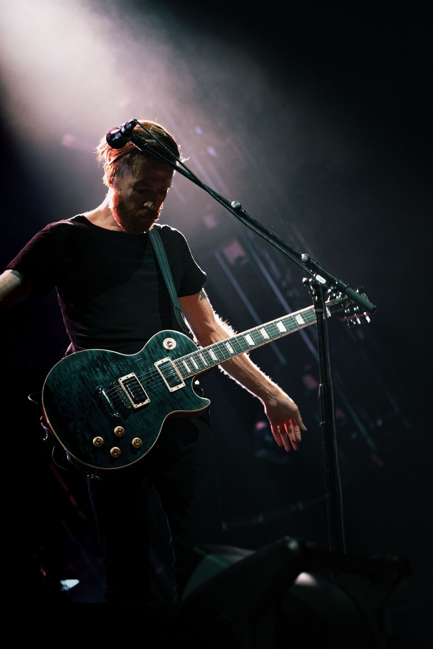 Guitarist playing black Les Paul guitar on dimly lit concert stage.