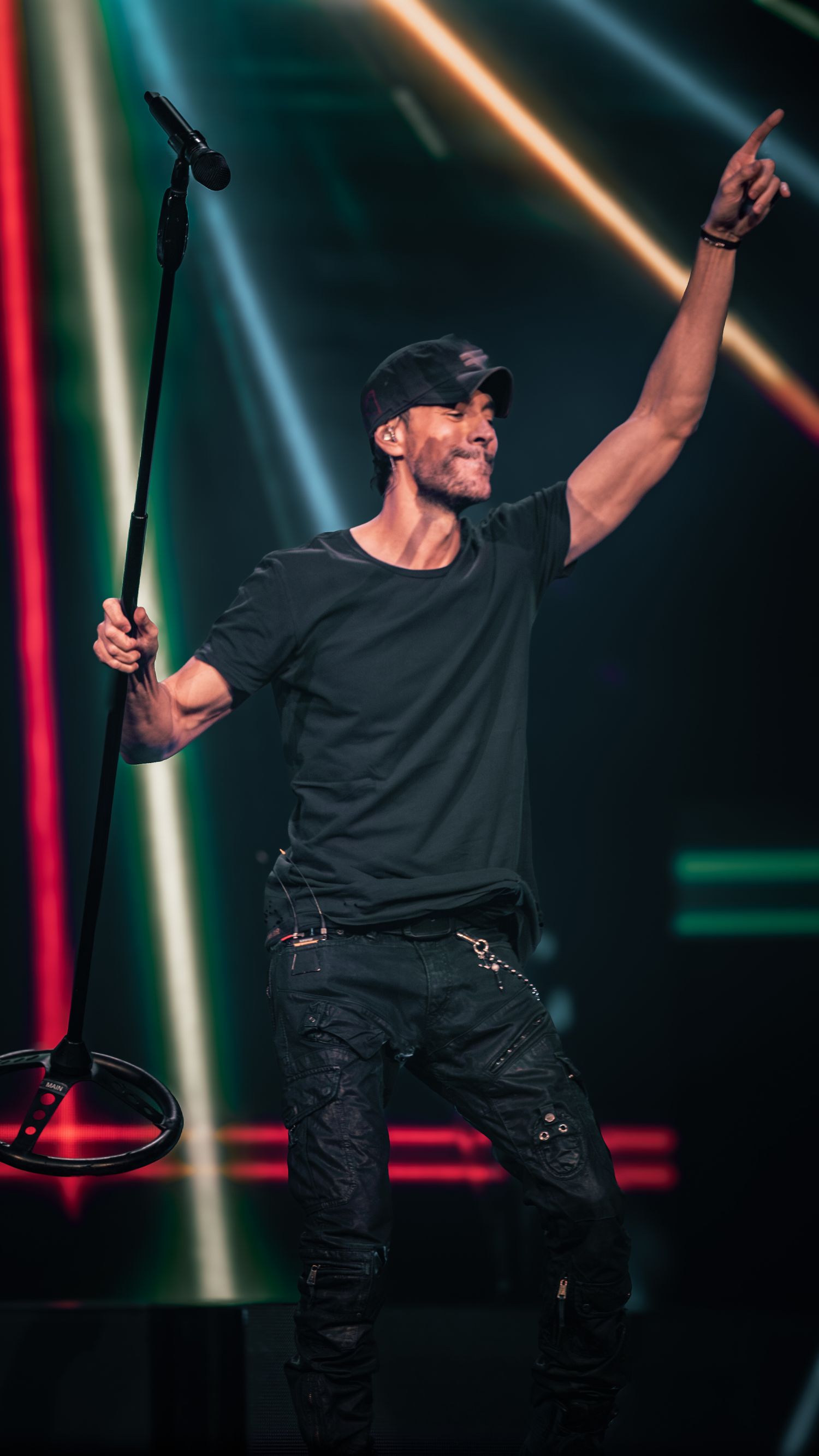 Energetic performer in black outfit raises arm while singing during concert with colorful light display.