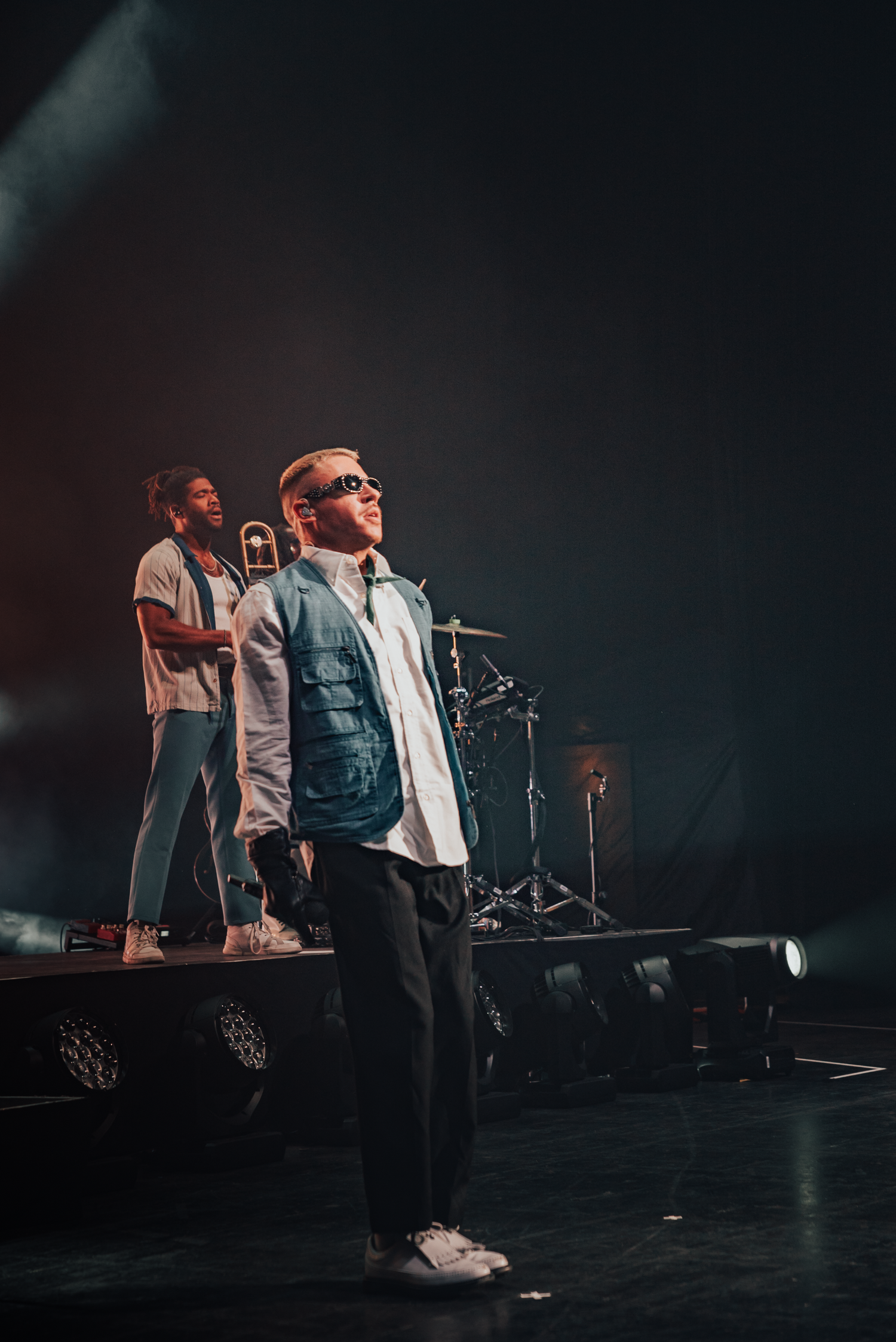 Stage performer in casual attire stands confidently under dramatic lighting during a live show.
