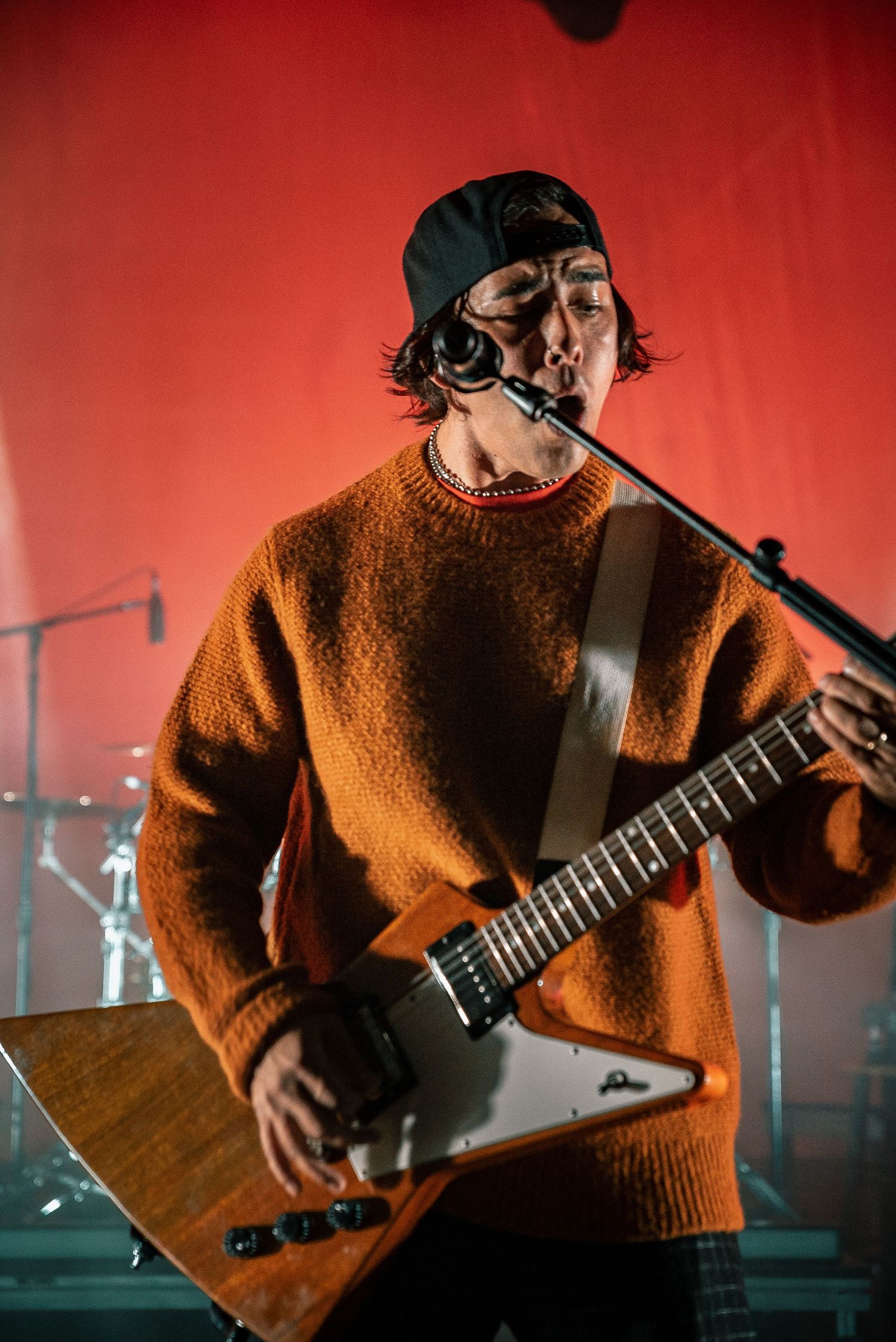 Musician in brown sweater plays guitar during live performance with red stage backdrop.