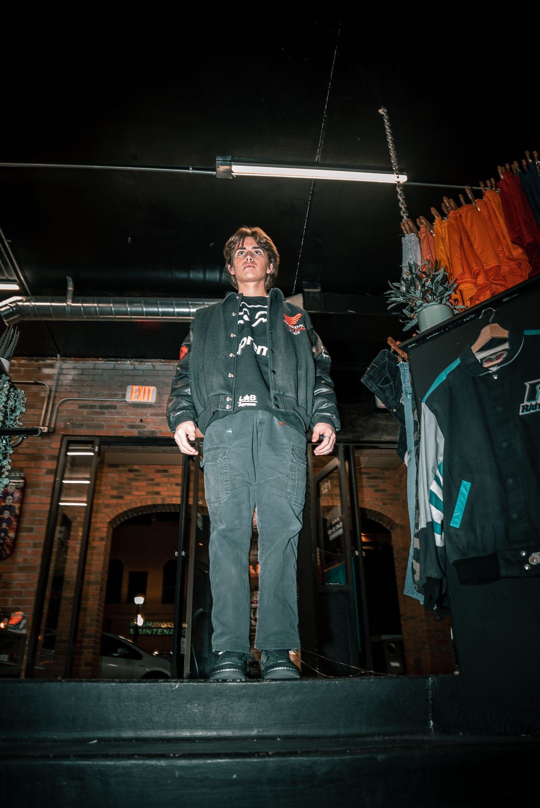 Figure standing on dark stage with hanging clothes and industrial lighting in background.