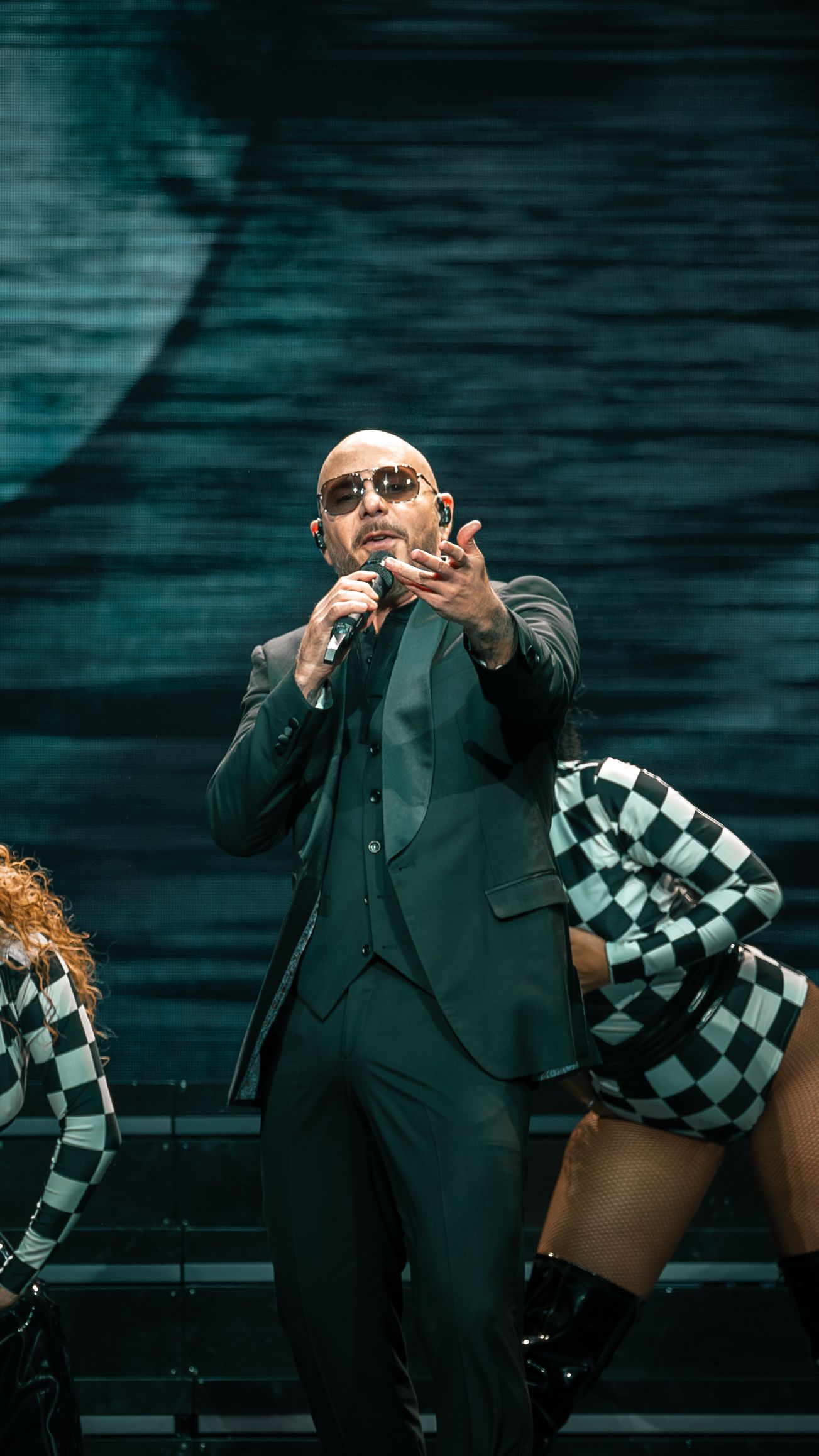Performer in green suit gestures dramatically on stage with dancers in checkered outfits performing in background.