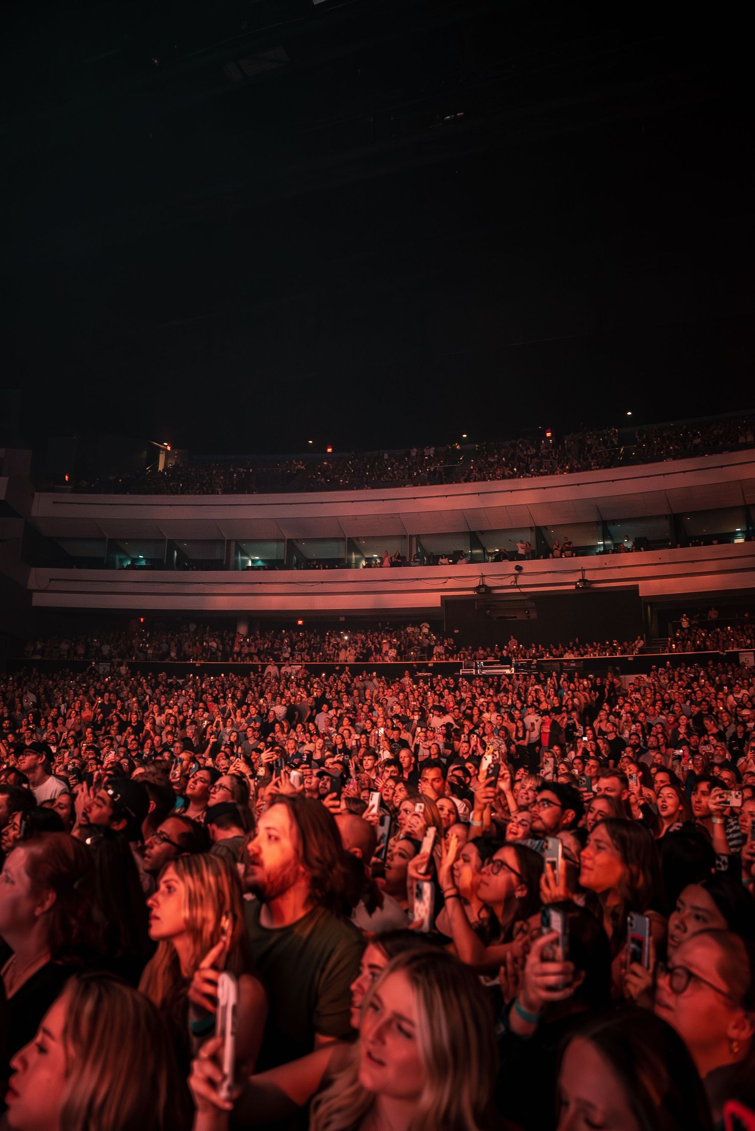 Large concert crowd illuminated by red stage lights filling an arena venue.