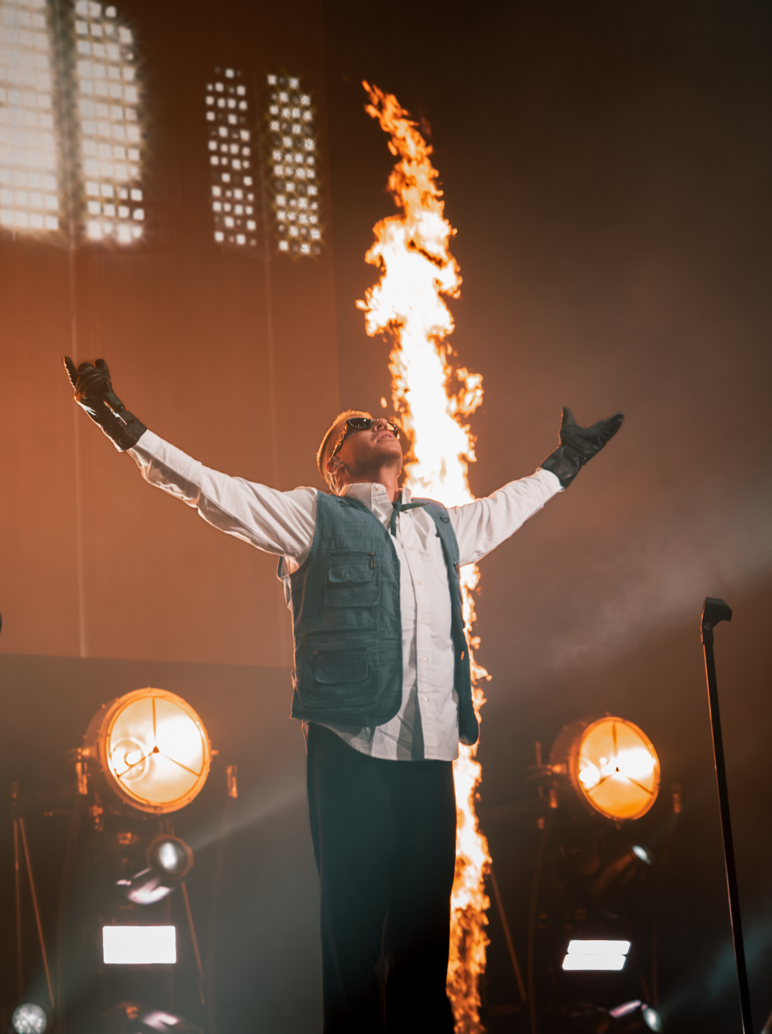 Concert stage with performer in vest silhouetted against dramatic fire effects.