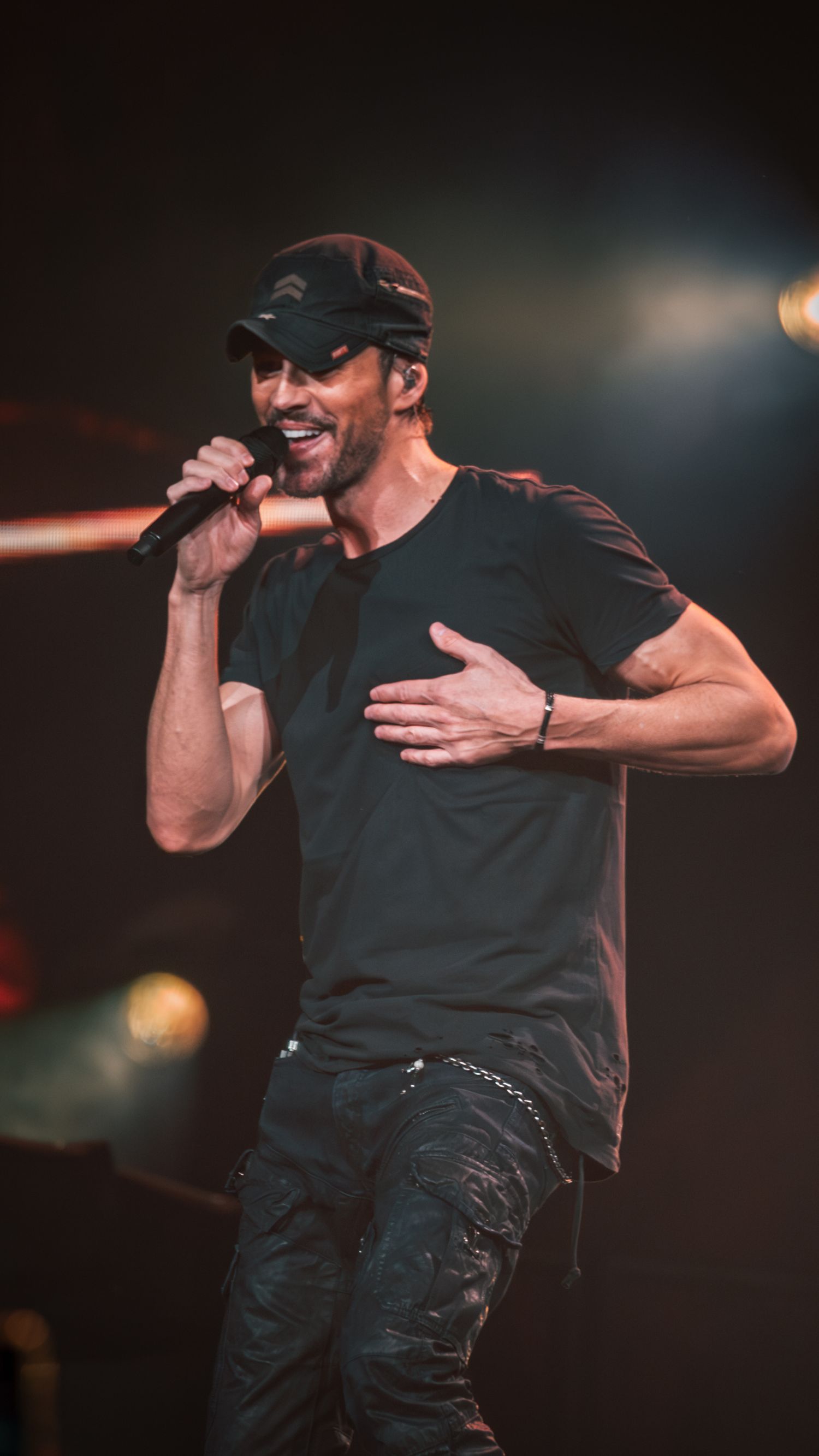 Male performer in black t-shirt and baseball cap singing emotively on stage with dramatic lighting effects.