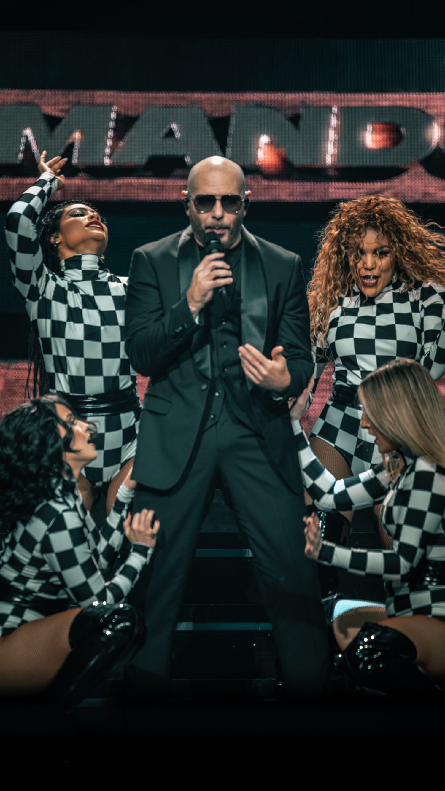 Energetic stage performance with dancers in black and white checkered outfits surrounding performer in dark suit.