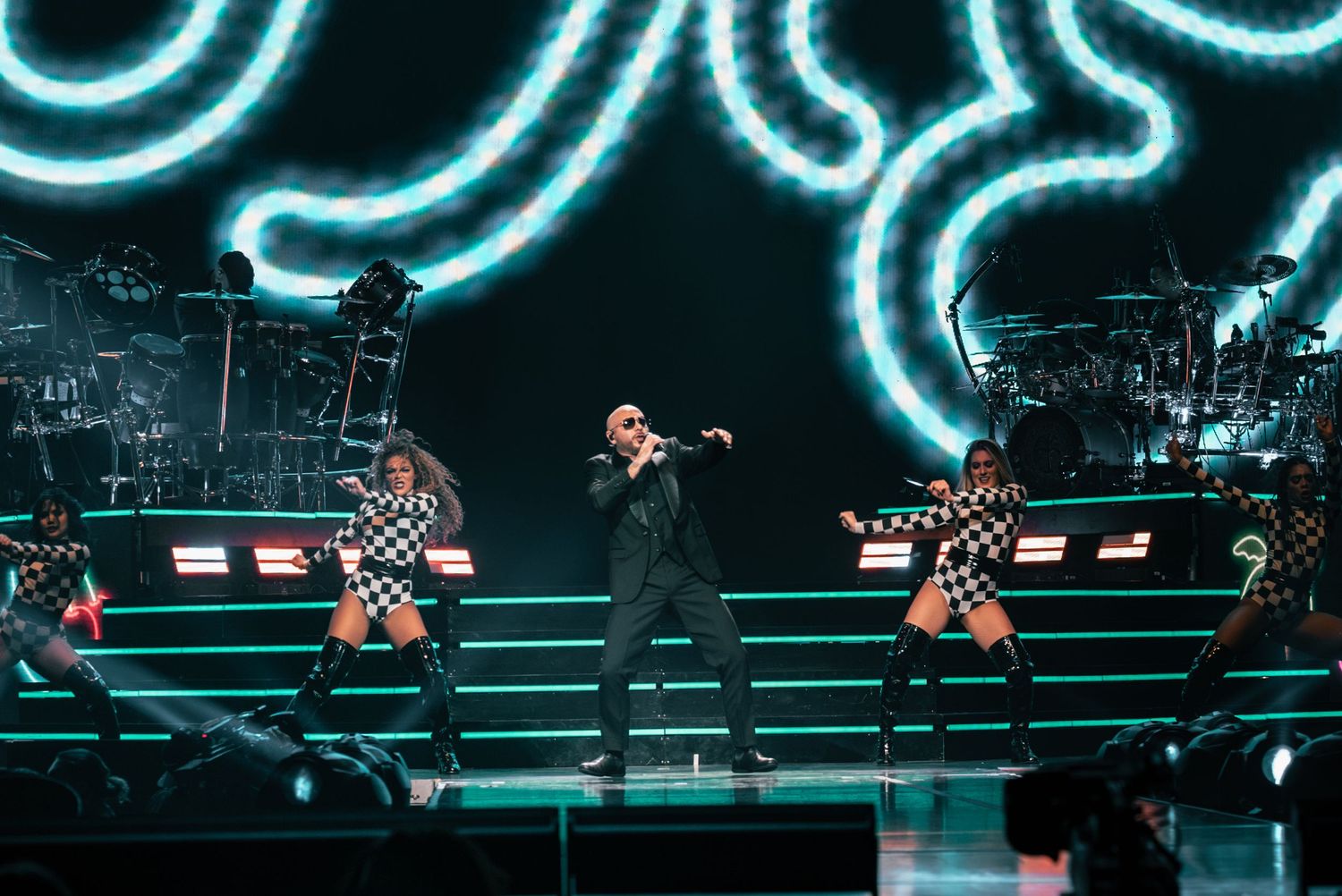 Stage performance with performers in black and checkered outfits against blue neon lighting display.