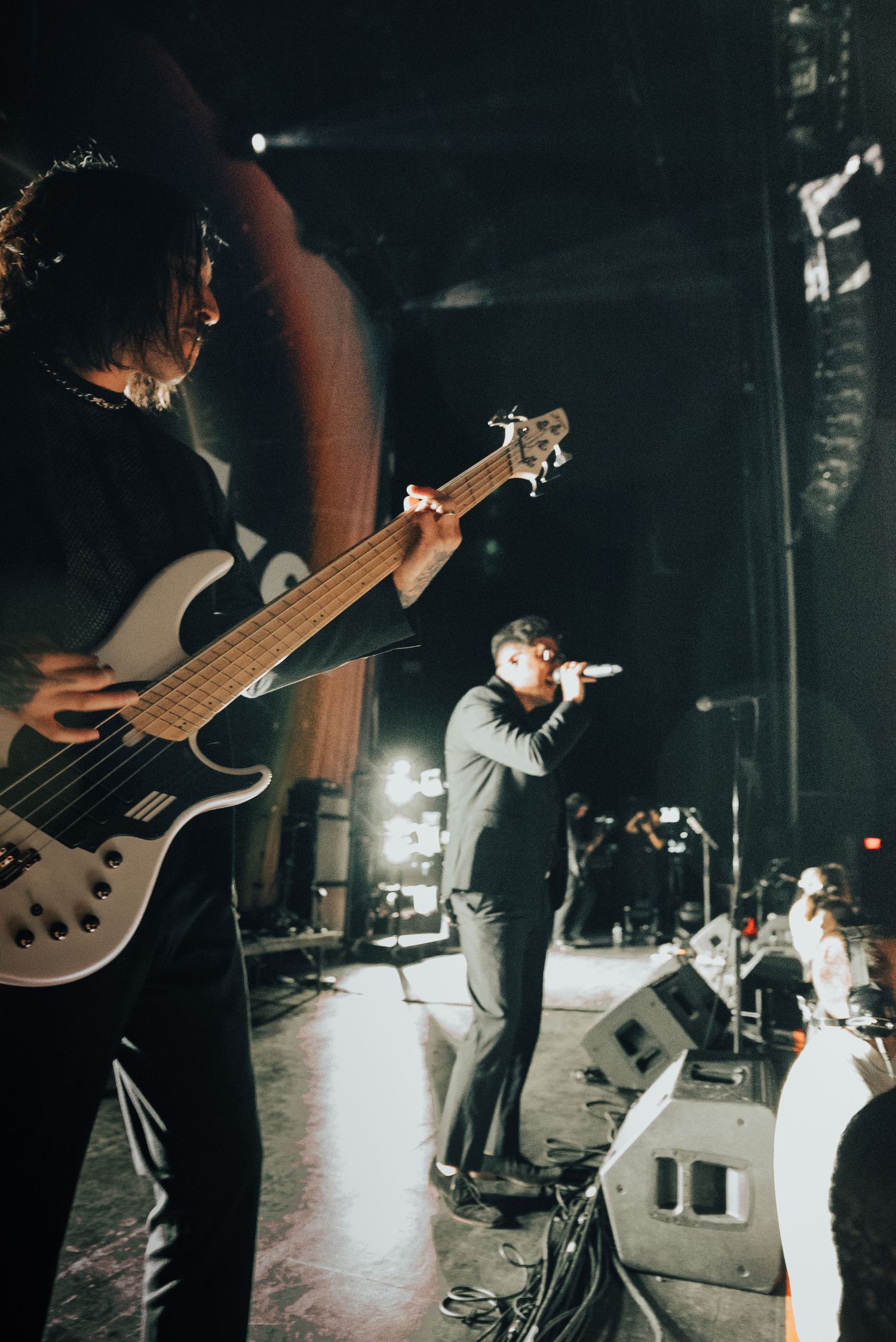 Rock band performs energetically on stage with bassist in foreground and vocalist at microphone.