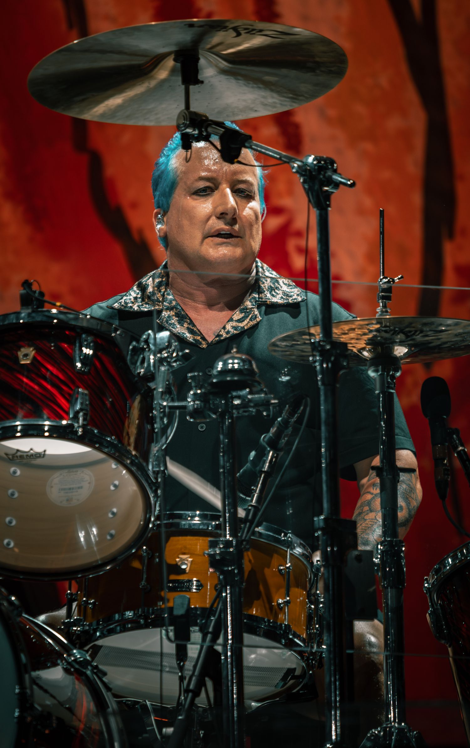 Drummer performs behind drum kit with cymbals and red stage lighting.