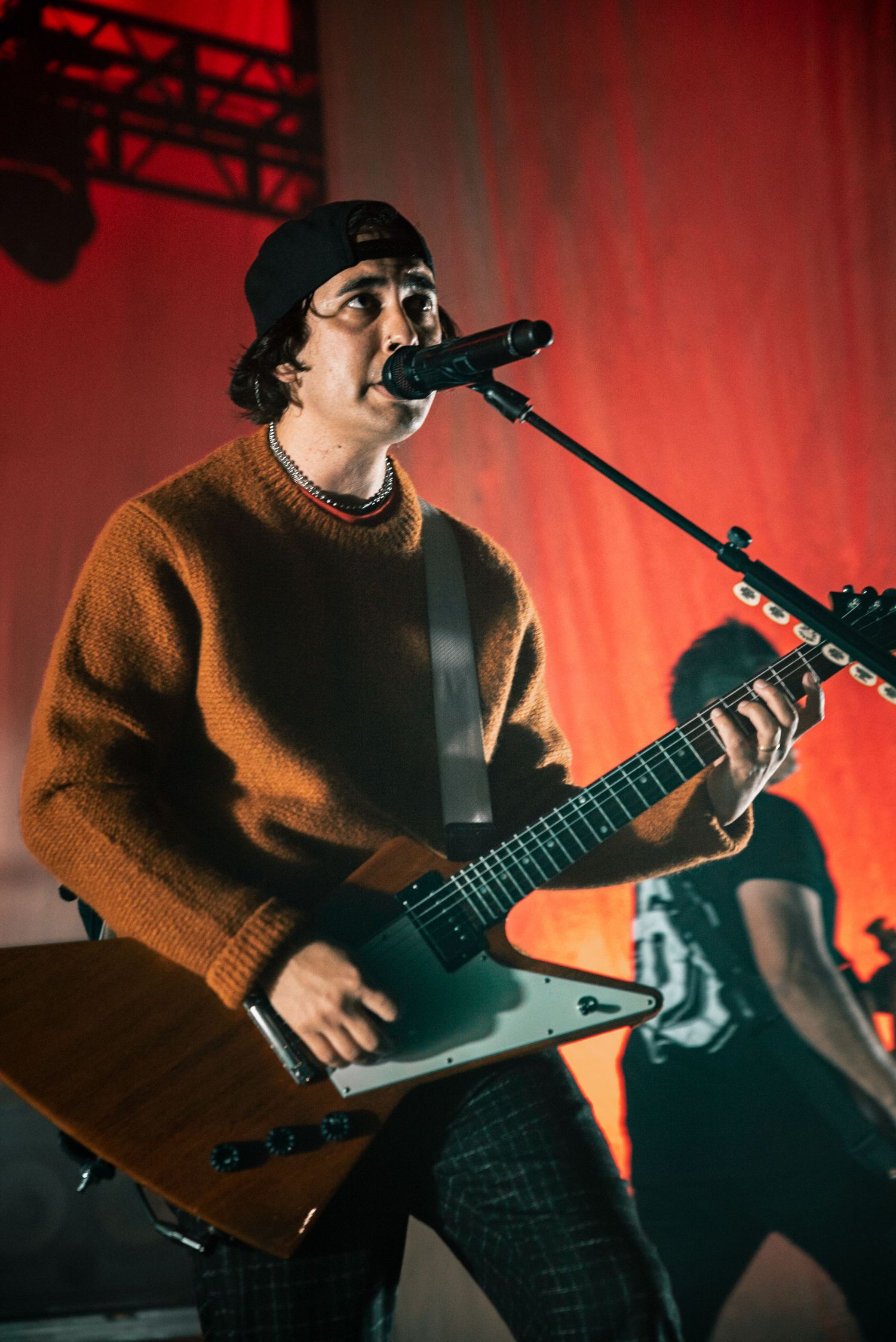Musician singing into microphone while playing metallic guitar against vibrant red stage backdrop.