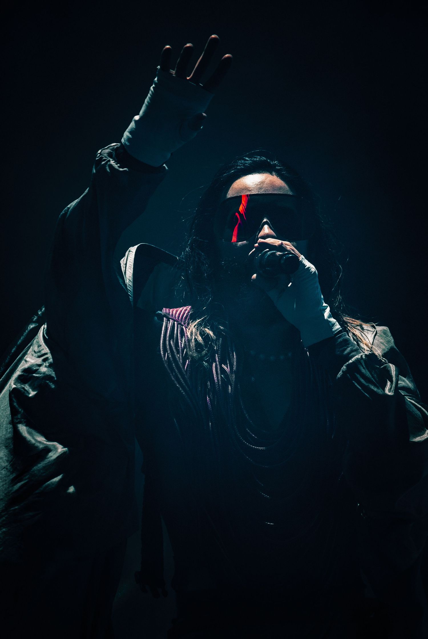 A dramatic concert photo showing a performer in dark clothing with red accents against black background.