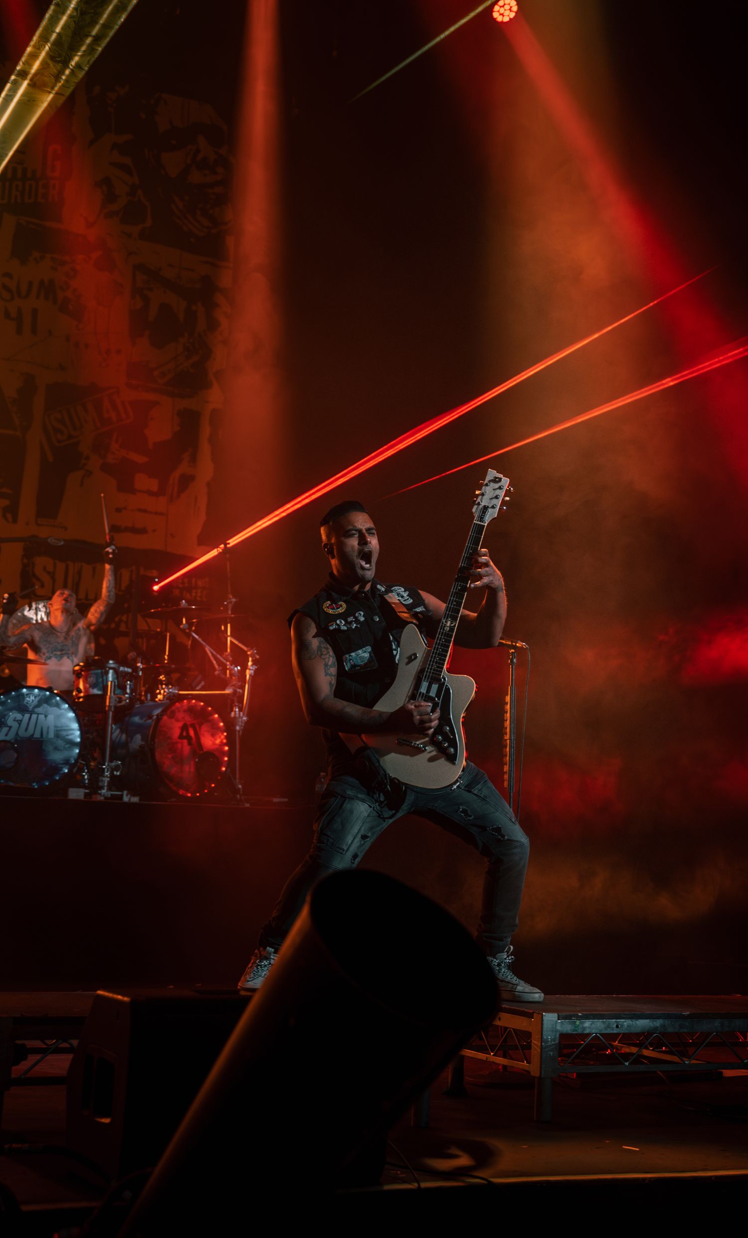 Guitarist performs on elevated platform with dramatic red stage lighting and laser beams.