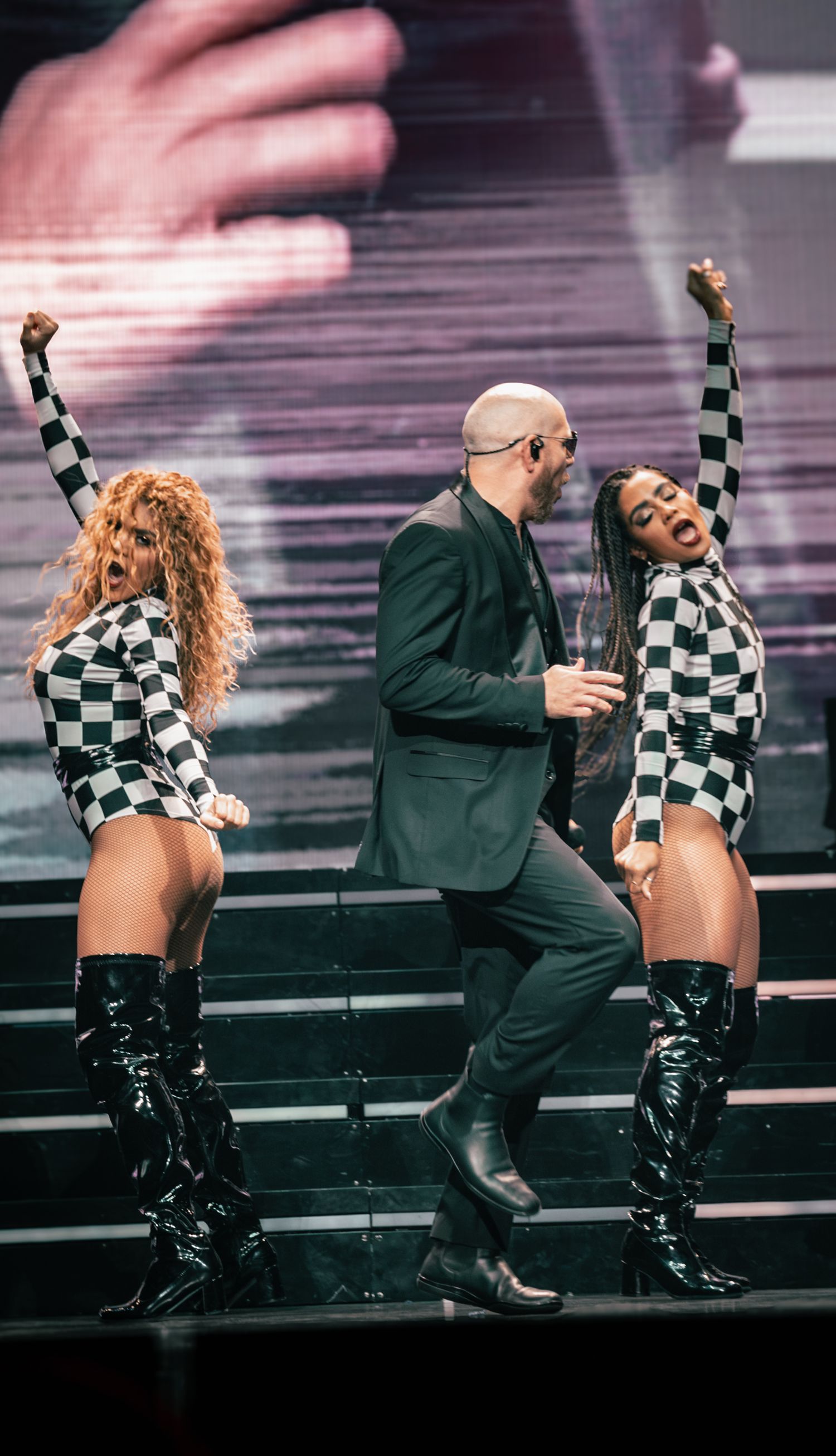 Dancers in checkered costumes perform alongside suited performer on concert stage steps.