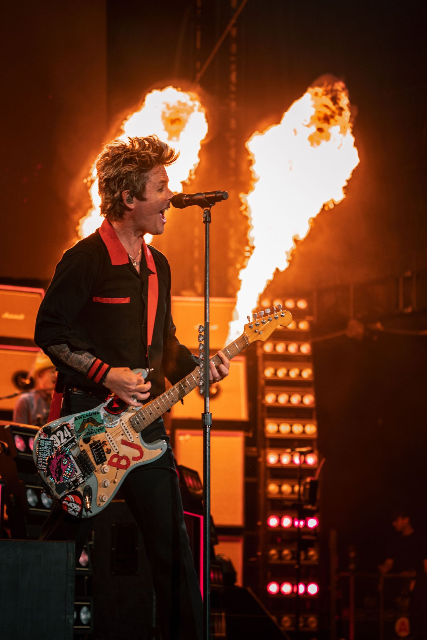 An energetic punk rock performance on stage with dramatic flame effects and colorful lighting behind the guitarist.