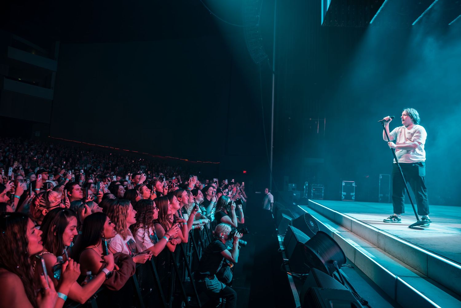 Concert performer singing passionately on stage with dramatic blue lighting and crowd visible in background.