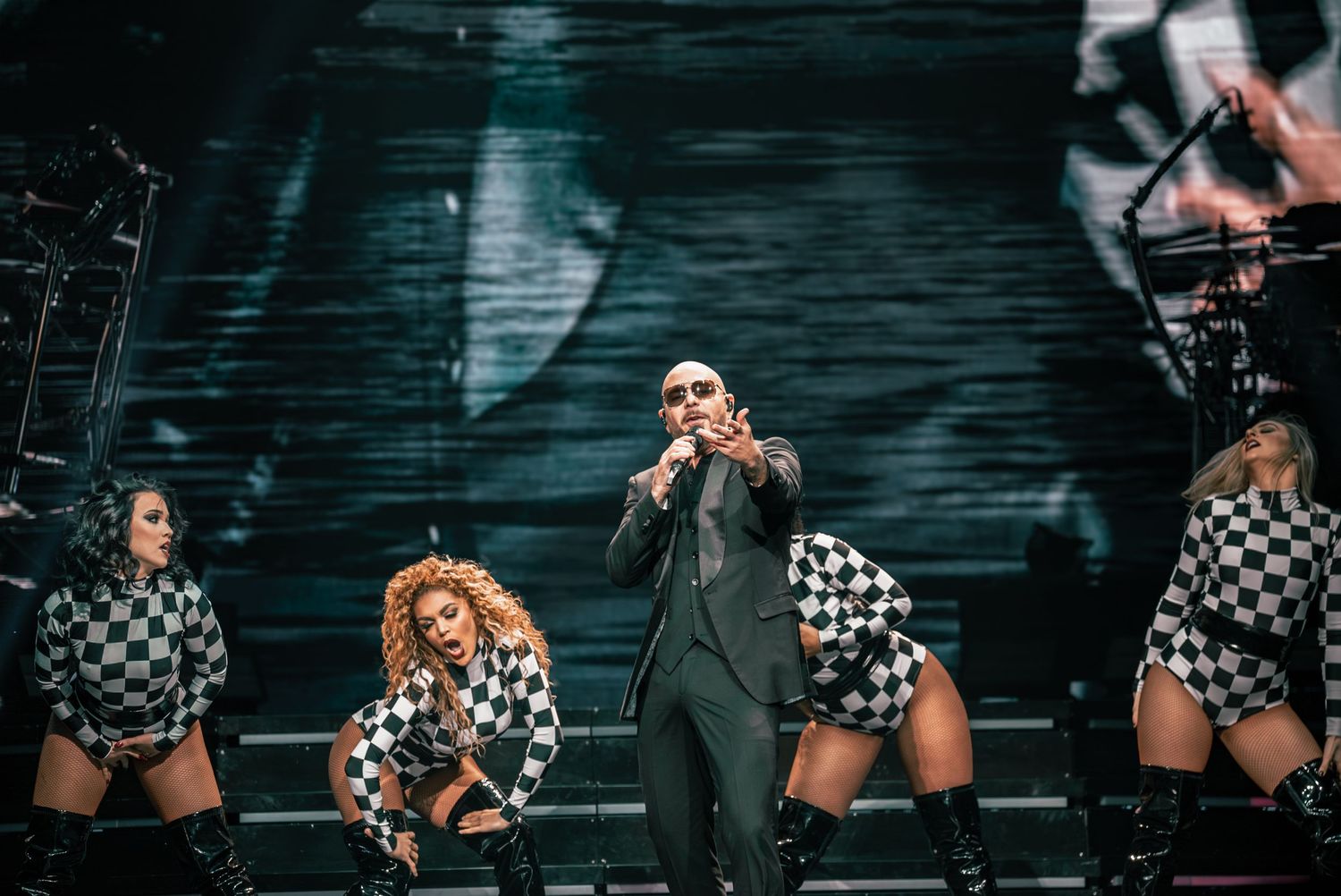 Dancers in checkered costumes perform alongside suited figure in dramatic stage production.