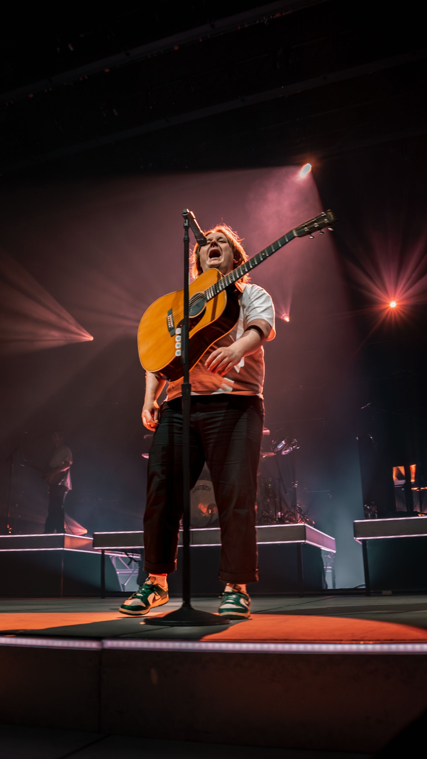 Performer with an acoustic guitar on a dramatically lit concert stage.