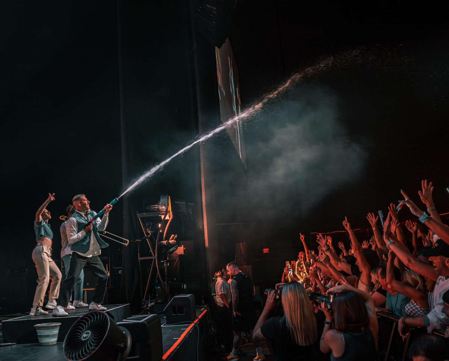 Concert crowd raising hands with stage performers silhouetted against dramatic lighting.