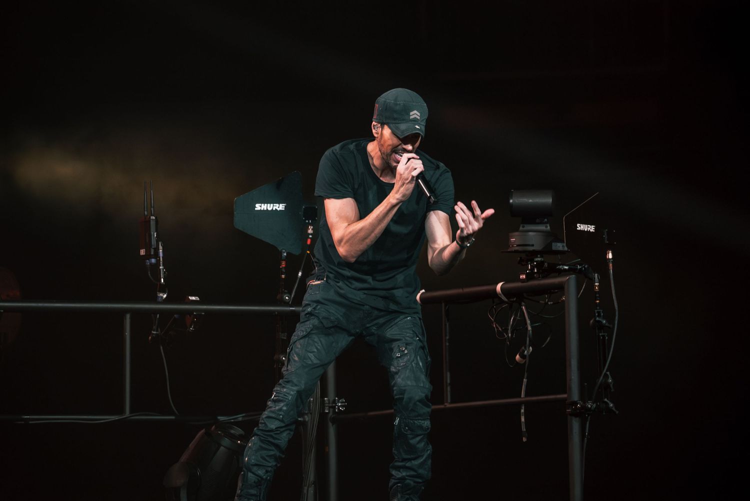 Concert performer in dark outfit sings intensely into microphone with stage equipment visible.