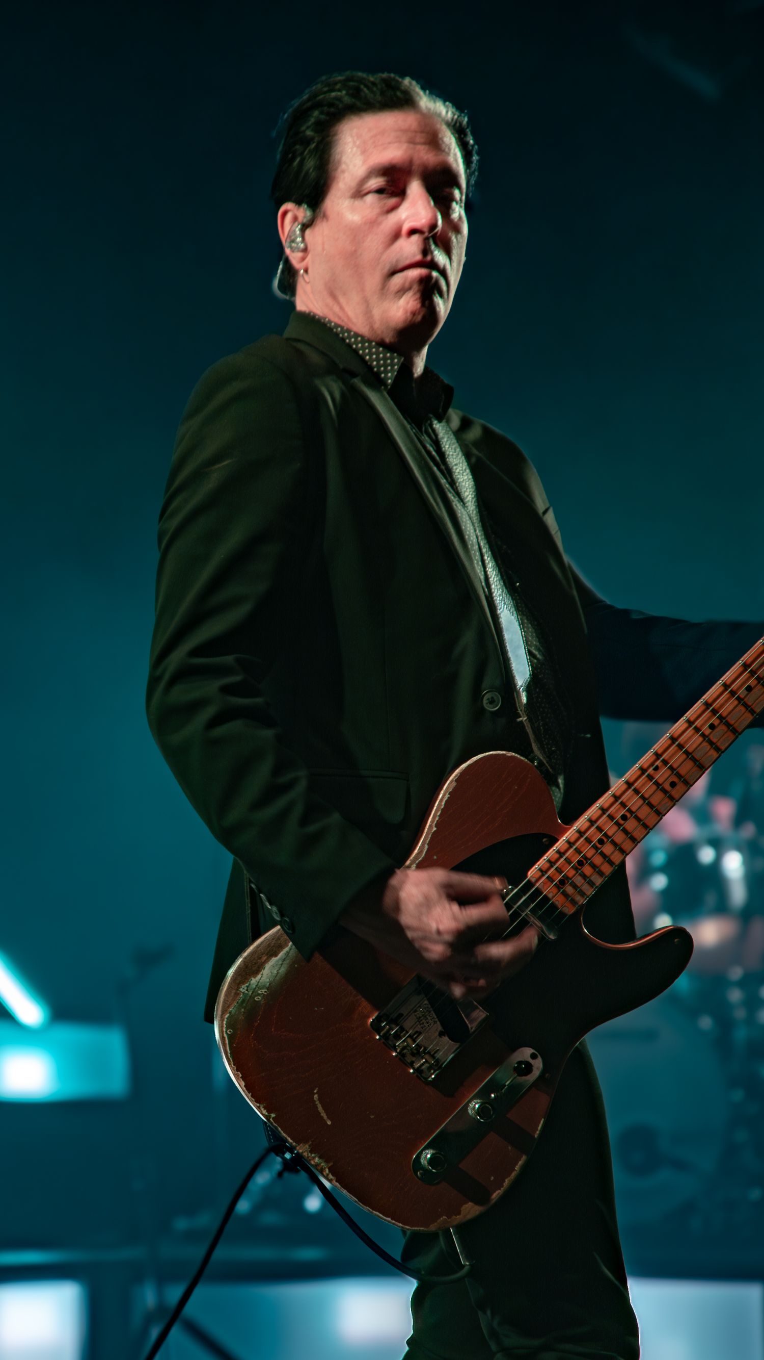 Performer in black suit plays acoustic guitar on dimly lit stage with blue lighting effects.