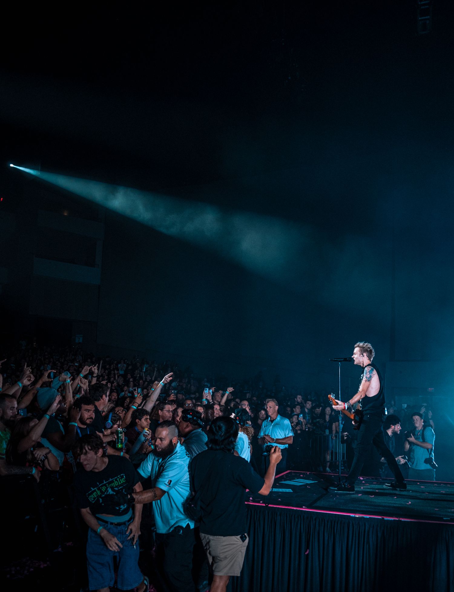 Enthusiastic concert crowd reaching towards performer on stage bathed in blue lighting.