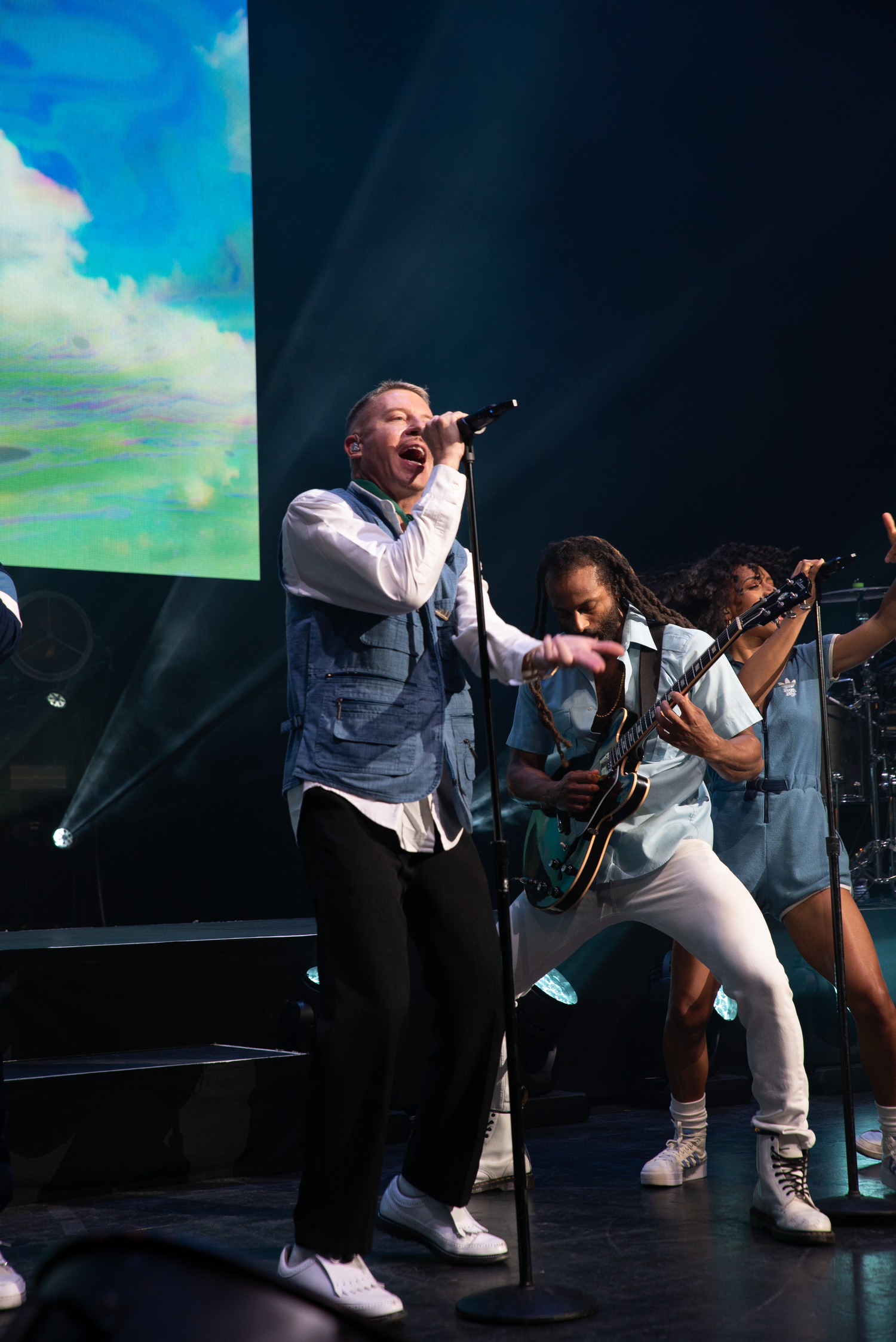 Musicians performing on stage with dramatic lighting and video screen backdrop during energetic concert performance.