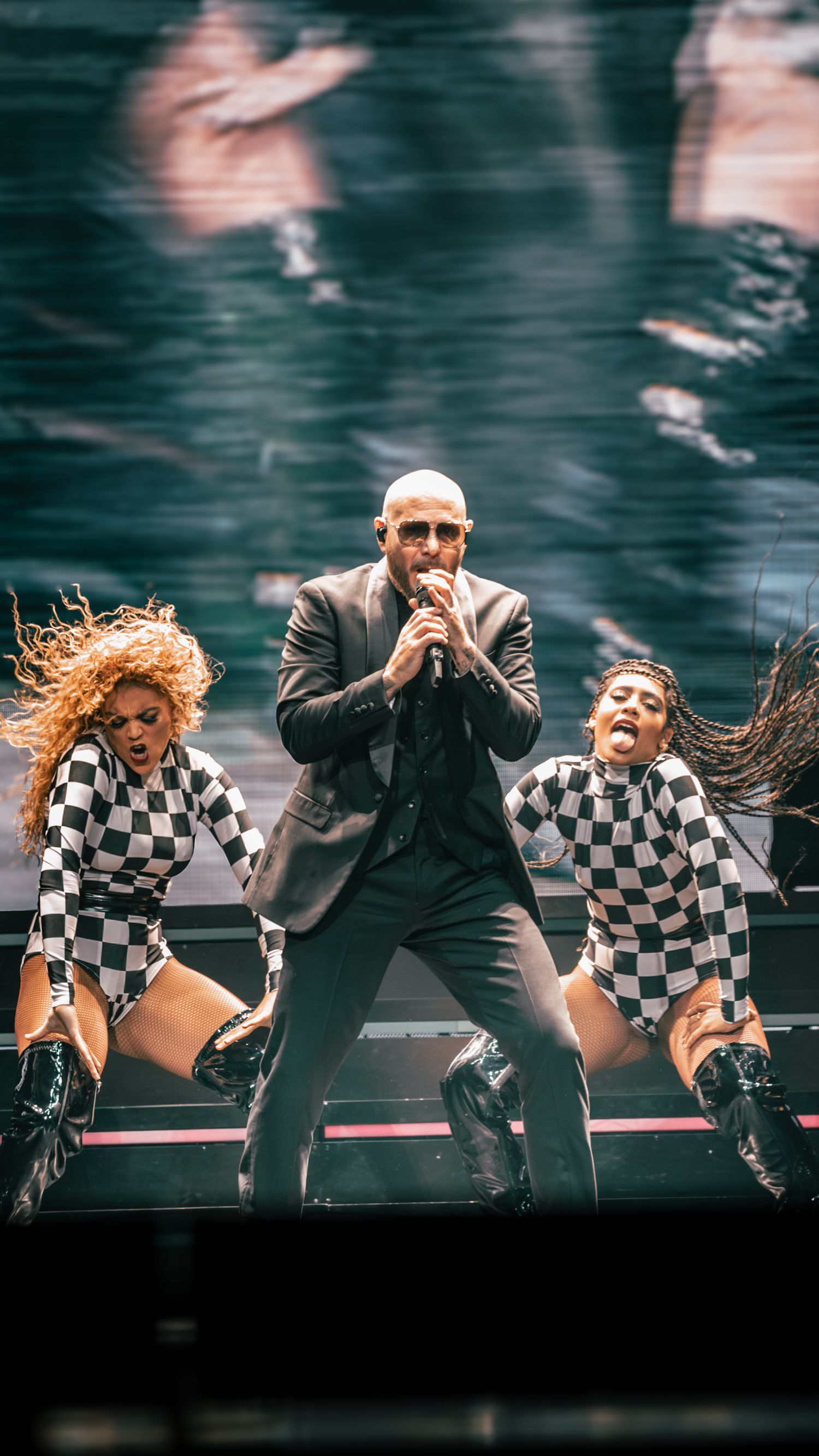 Dynamic concert scene with performers dancing in checkered outfits on illuminated stage.