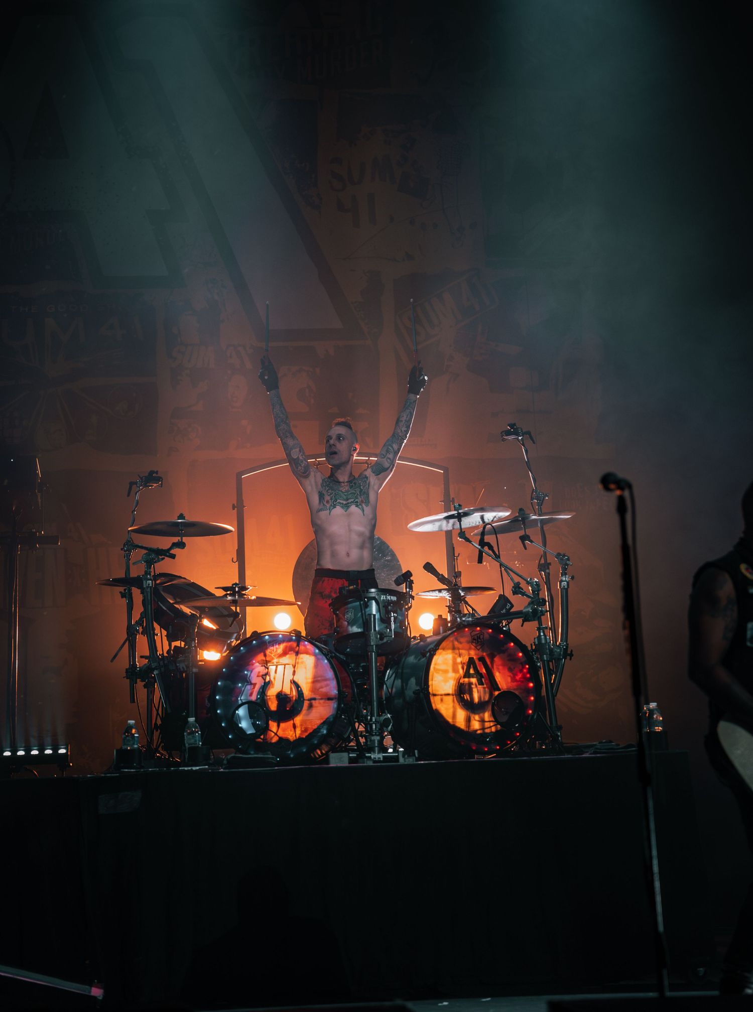 Drummer performs energetically behind lit drum kit with orange backlighting on stage.