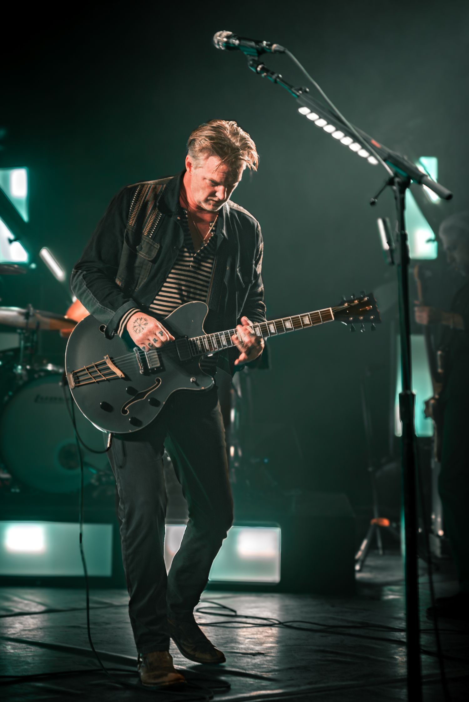 Musician crouches while playing electric guitar on darkened stage with green lighting effects.