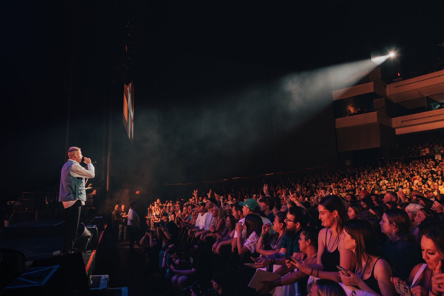 Concert venue filled with enthusiastic crowd watching performer on stage under dramatic lighting.