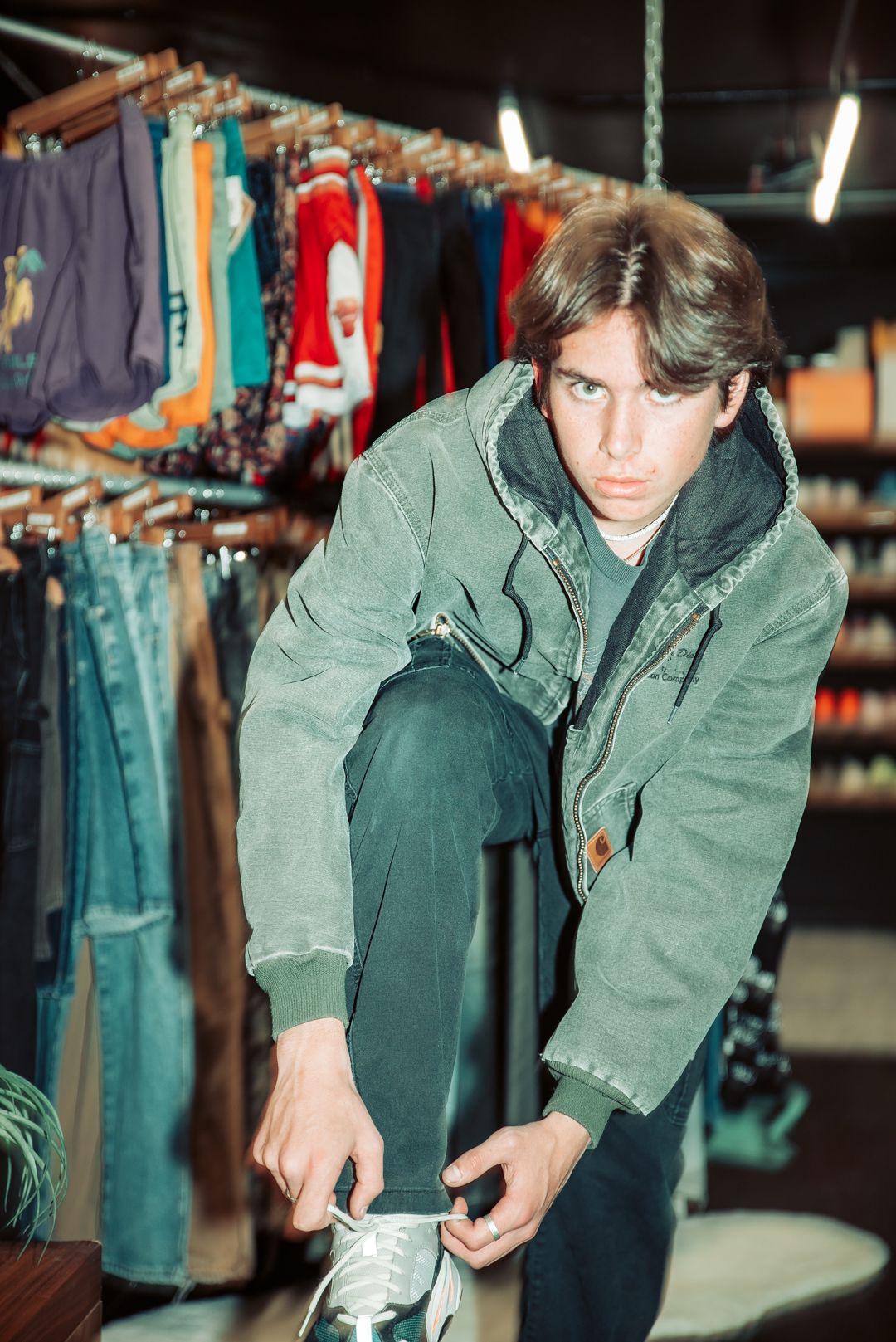 Figure in olive jacket tying shoes in clothing store with racks of garments.