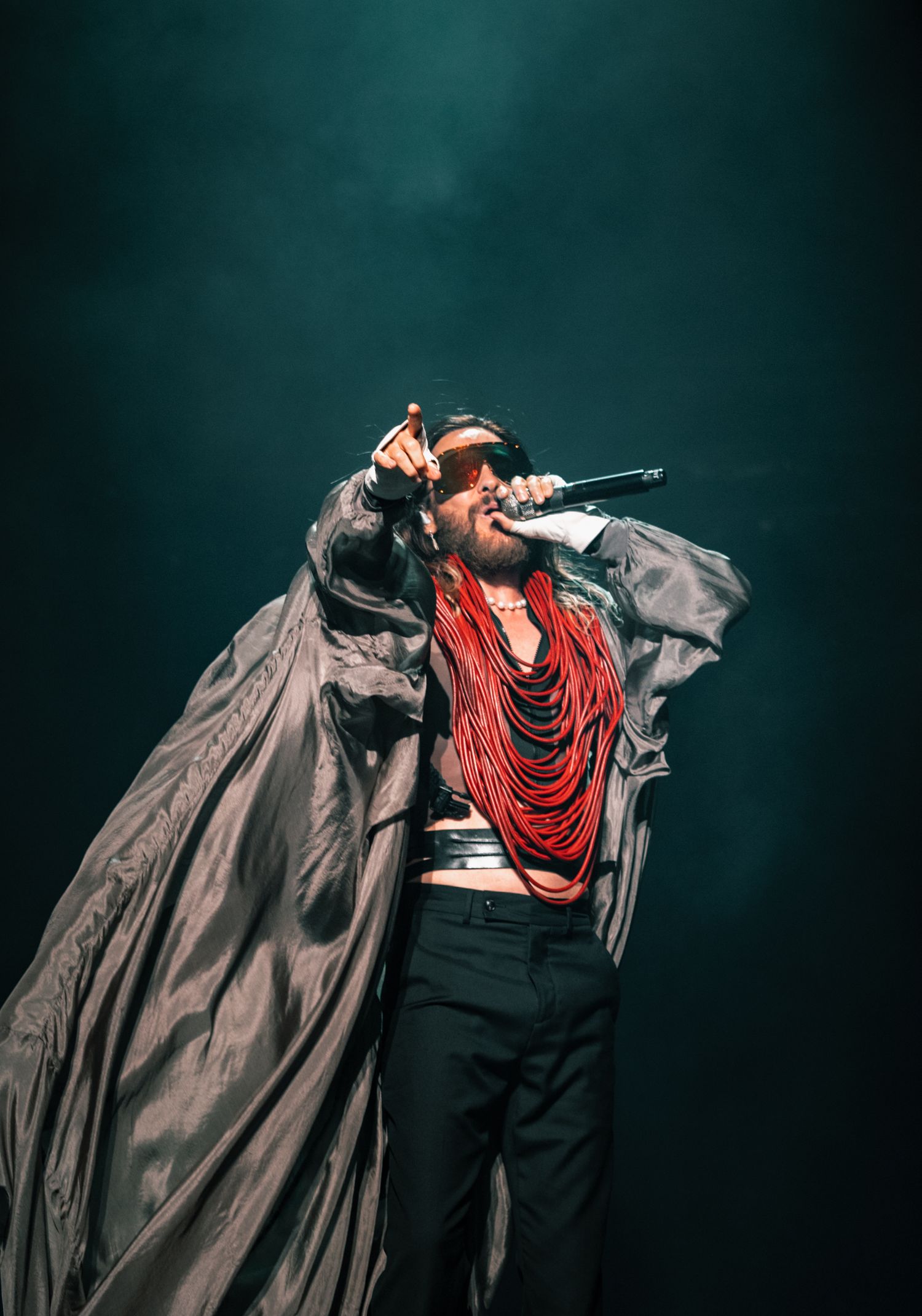 An energetic performer wearing flowing black coat and red necklaces singing passionately on stage.
