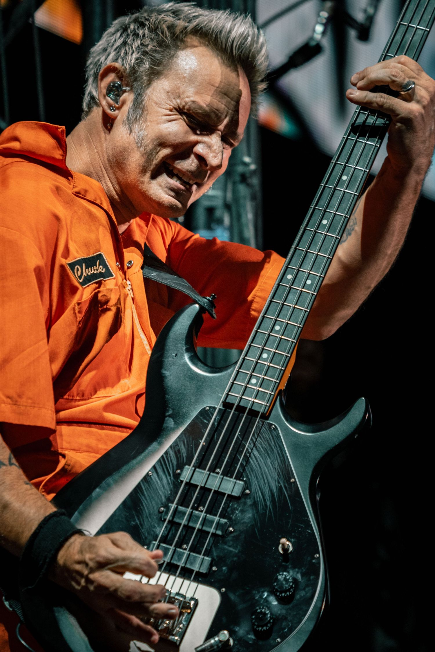 A bassist in orange workwear plays passionately on a dark electric bass guitar during a live performance.