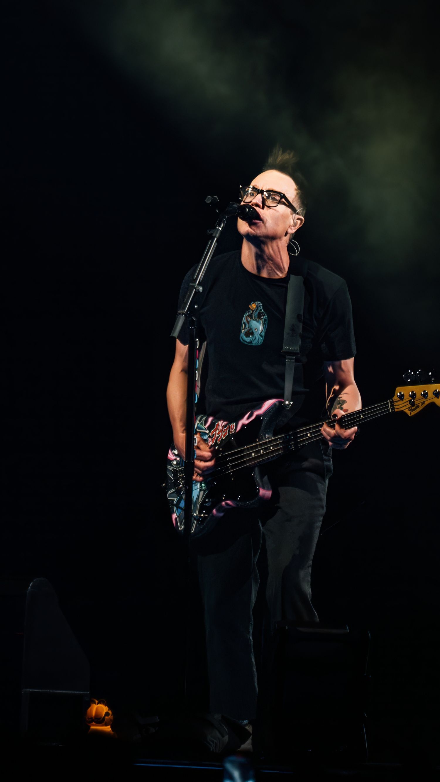 Bass player in black attire performs on darkened stage with dramatic lighting.