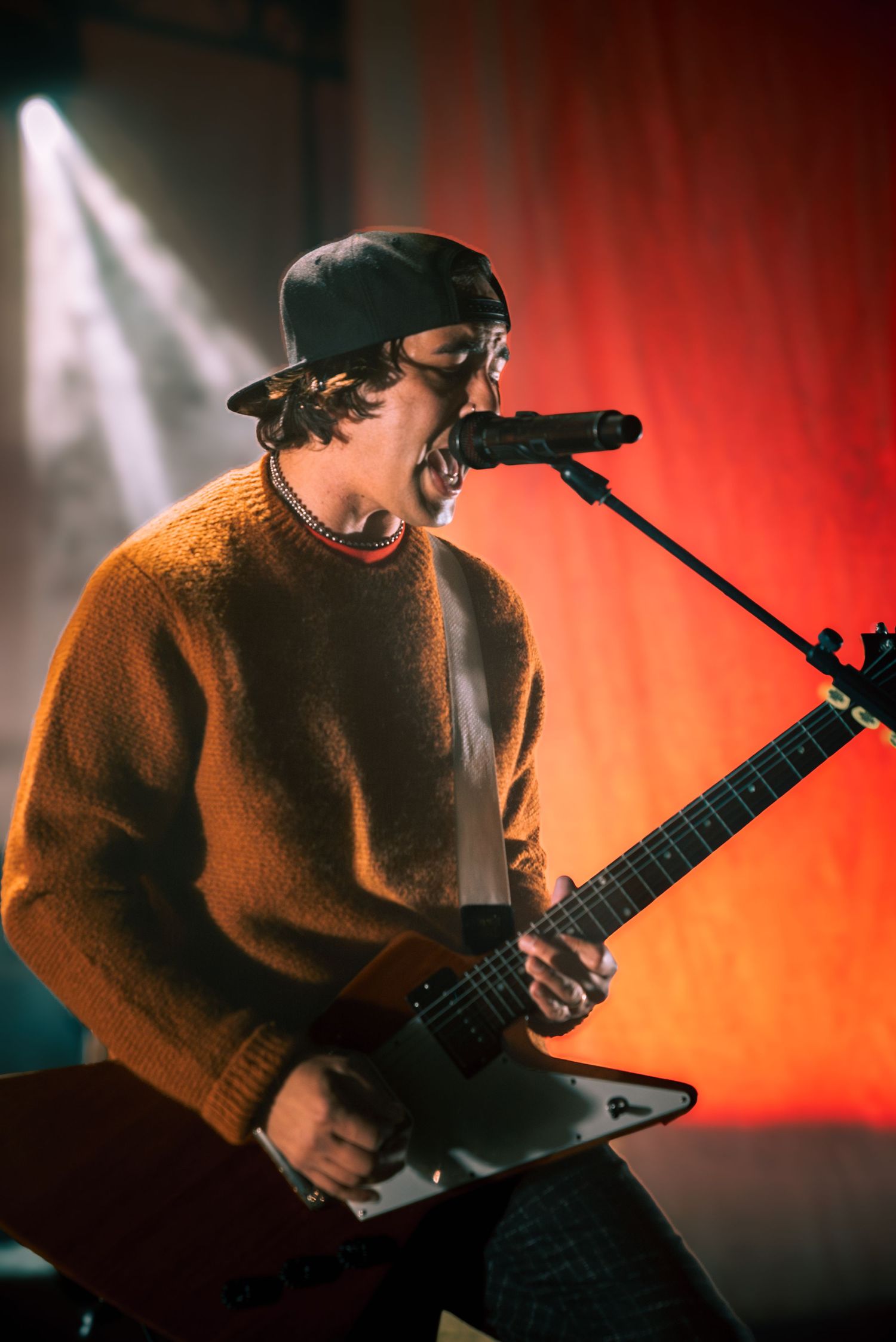 Performer in brown sweater plays angular electric guitar while singing into microphone on dimly lit stage.